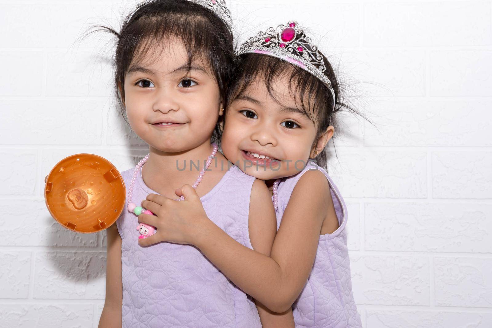 Female asian identical twins sitting on chair with white background. Wearing purple dress and accessories. Hugging each other while standing by billroque