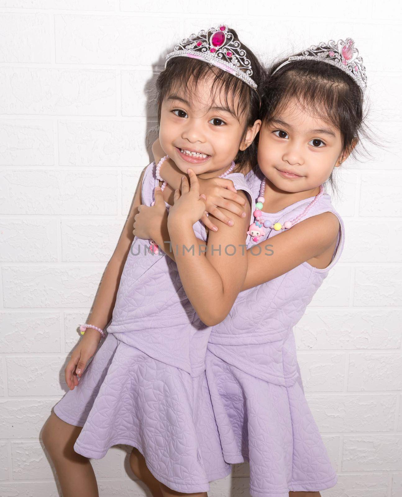 Female asian identical twins sitting on chair with white background. Wearing purple dress and accessories. Standing hugging each other and having fun