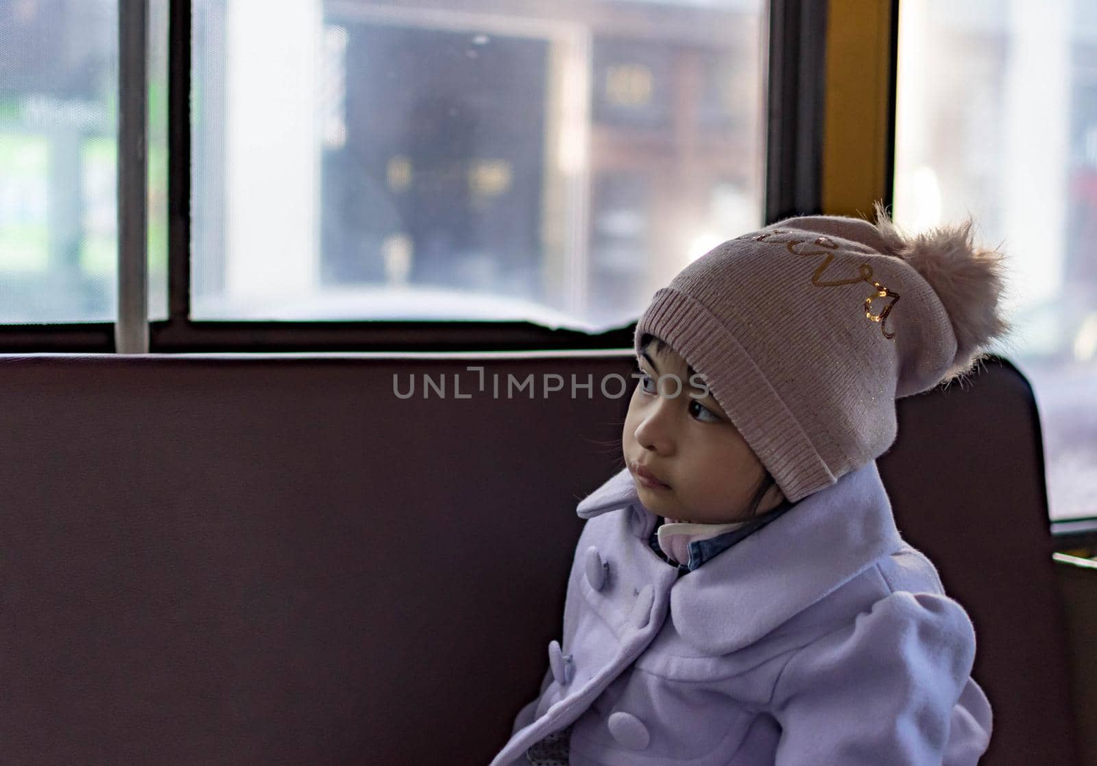 pretty female child wearing purple coat and cute bonnet while sitting inside a bus and enjoying the view from the window
