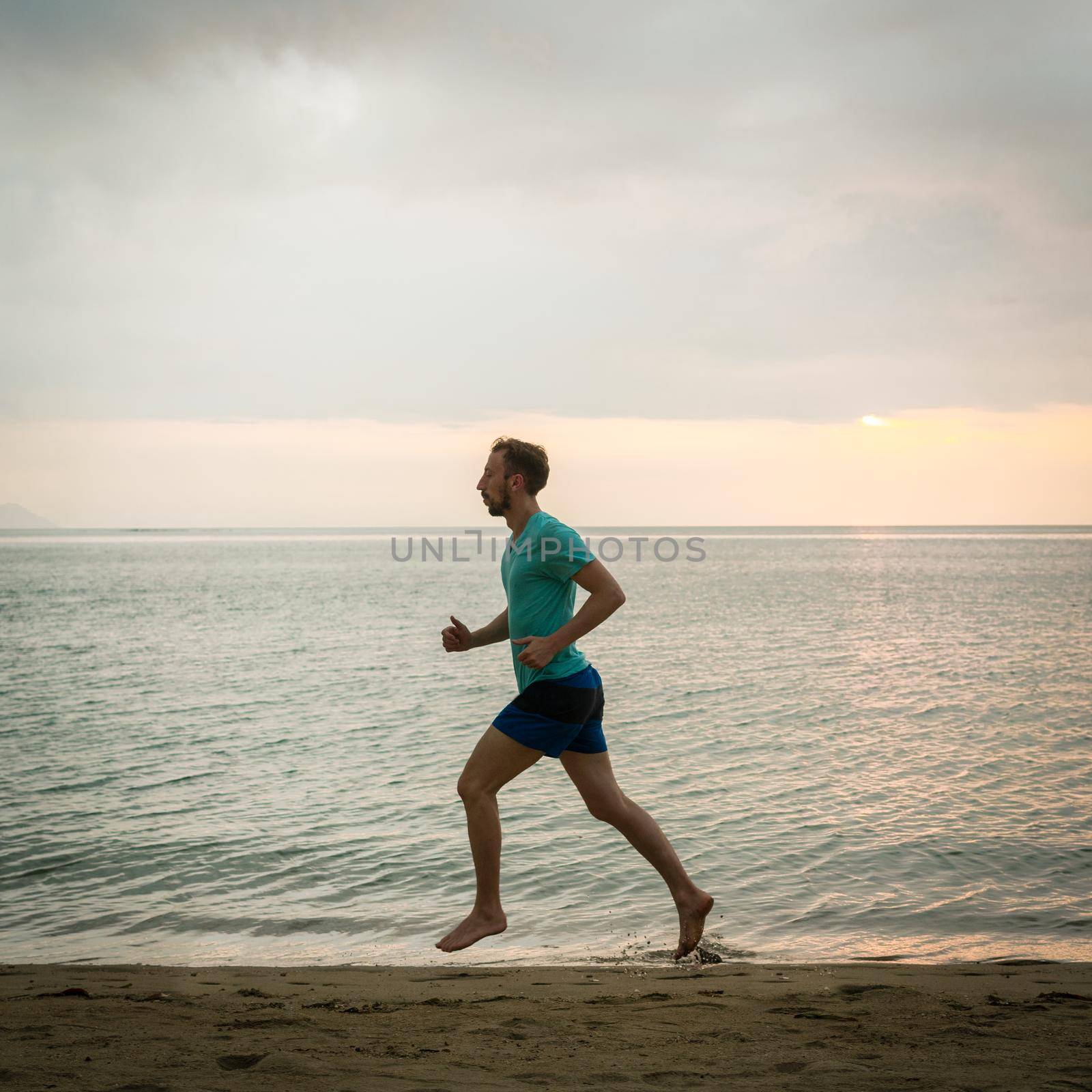 Athletic young man running on the beach during cardio workout session by Kzenon