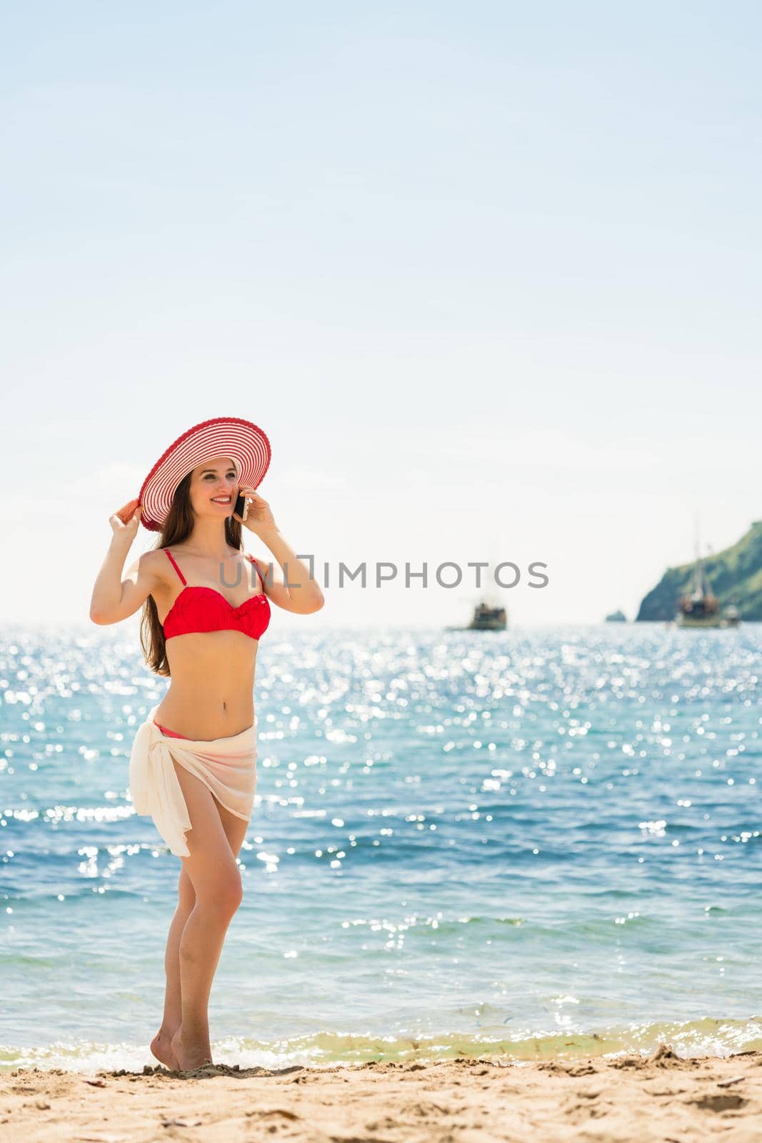 Fashionable fit young woman wearing red swimwear while talking on mobile phone on the beach during summer vacation in Flores Island, Indonesia