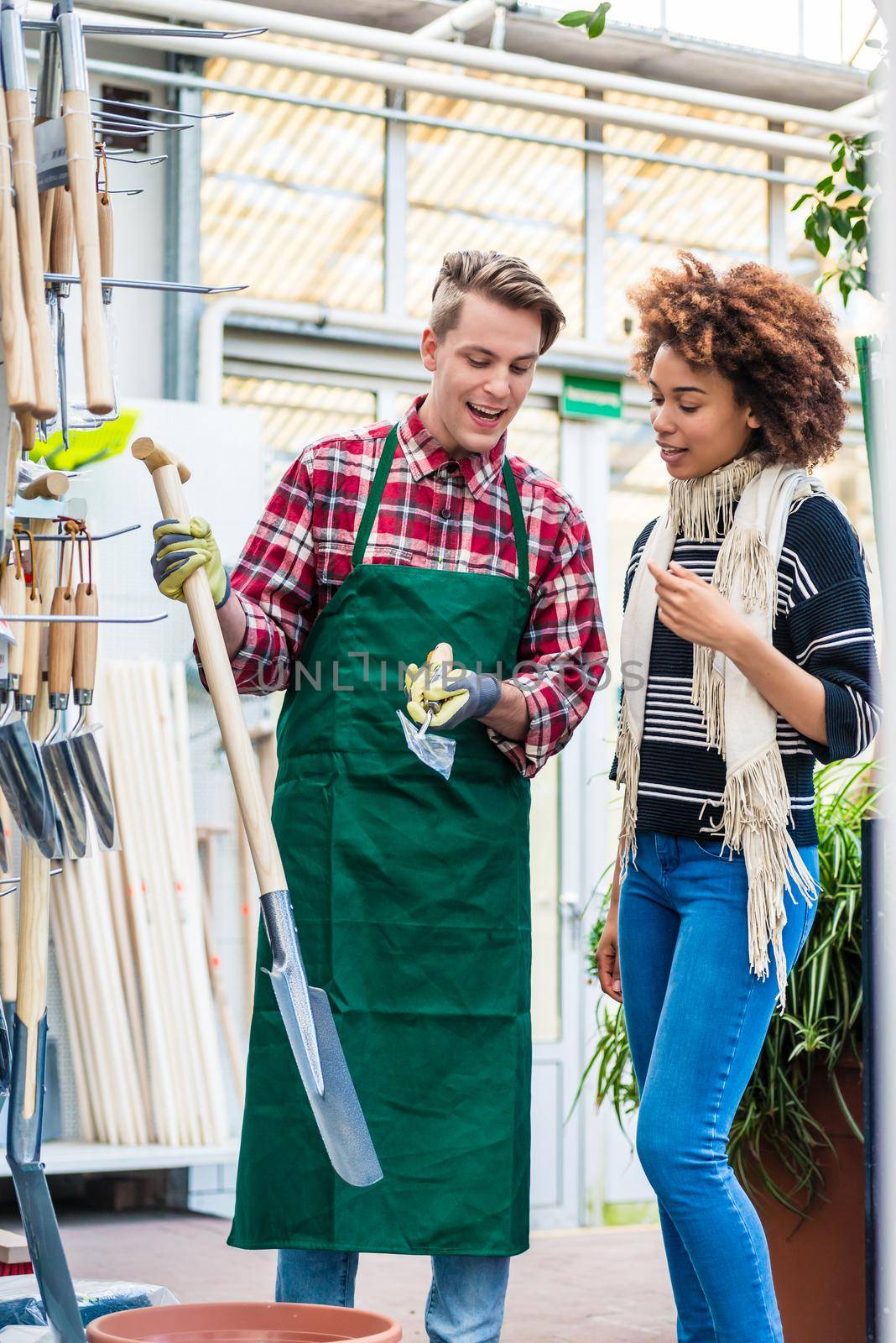 Handsome and cheerful worker wearing a green apron while helping a customer with choosing a gardening tool in a modern flower shop