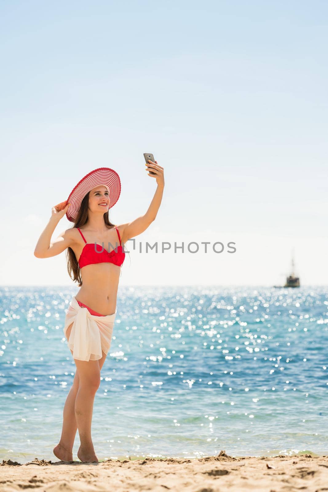 Beautiful young woman wearing a trendy striped hat while posing for a selfie picture on the beach during summer vacation in Flores Island, Indonesia