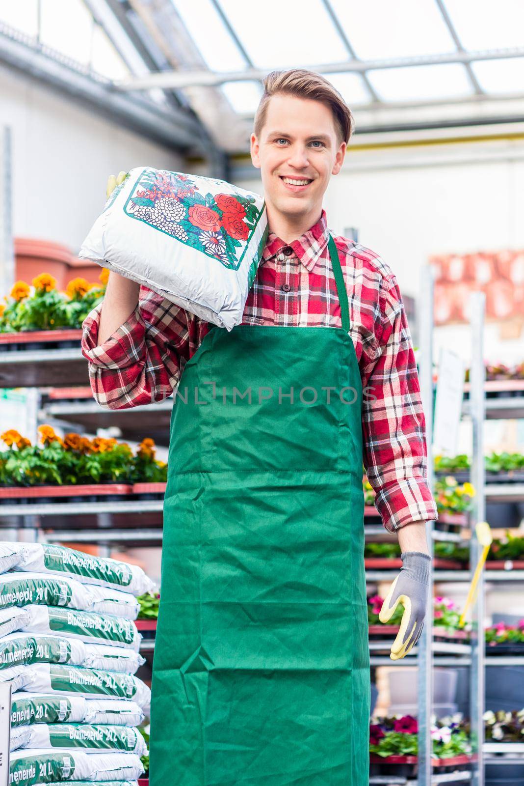 Cheerful young man carrying a bag of potting soil while working in a flower shop by Kzenon