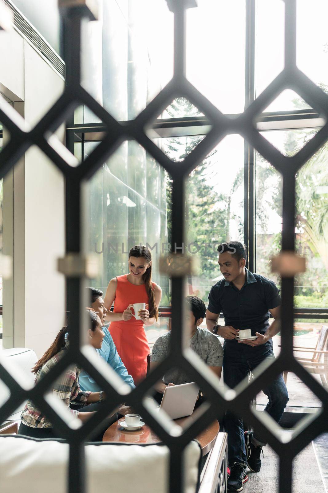 Full length of a multi-ethnic group of five cheerful employees drinking coffee, while watching a funny video on laptop during break in the lounge area of a modern multinational company