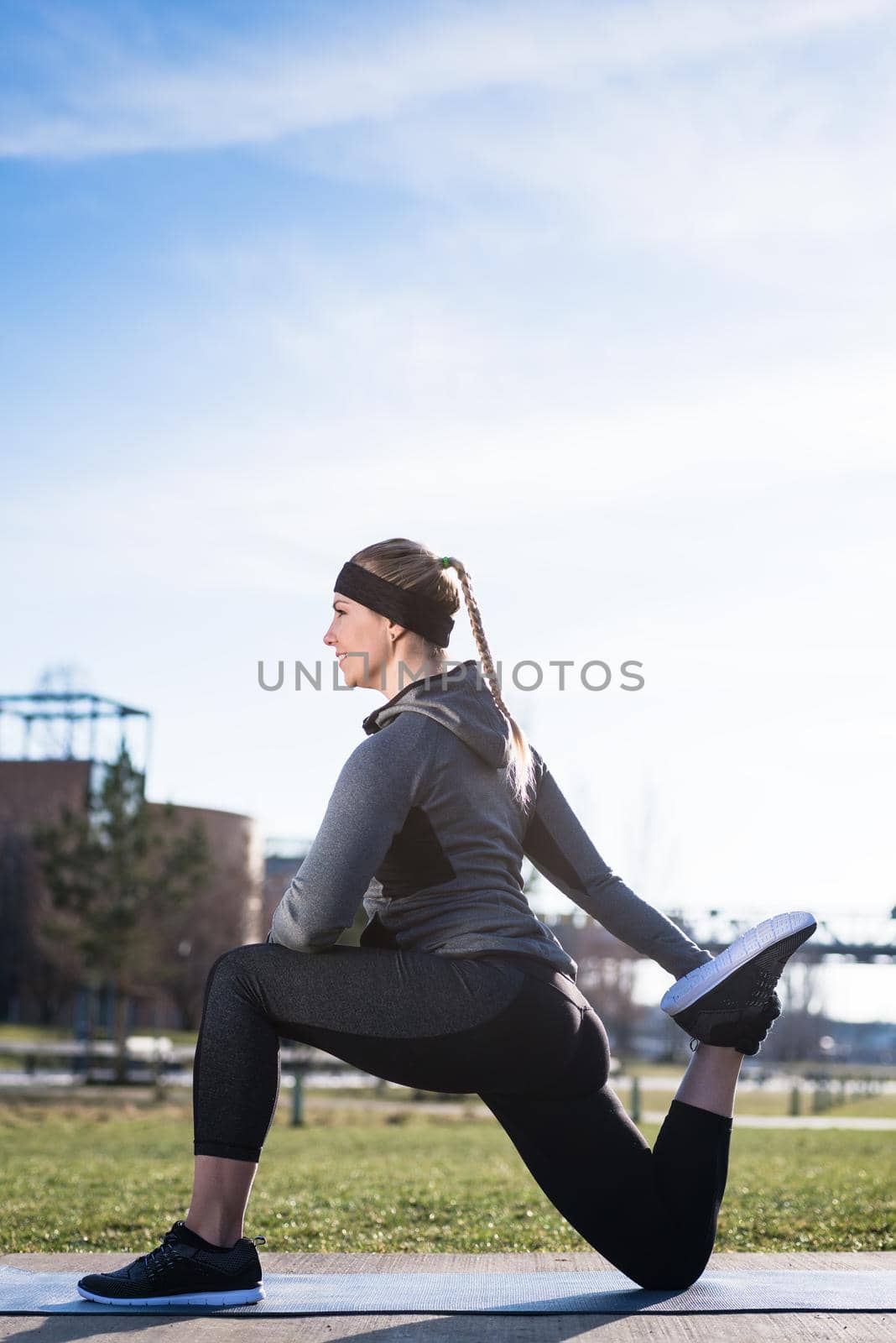 Young woman stretching her quadriceps muscles by grabbing her ankle during outdoor warming up routine in the park in a sunny day