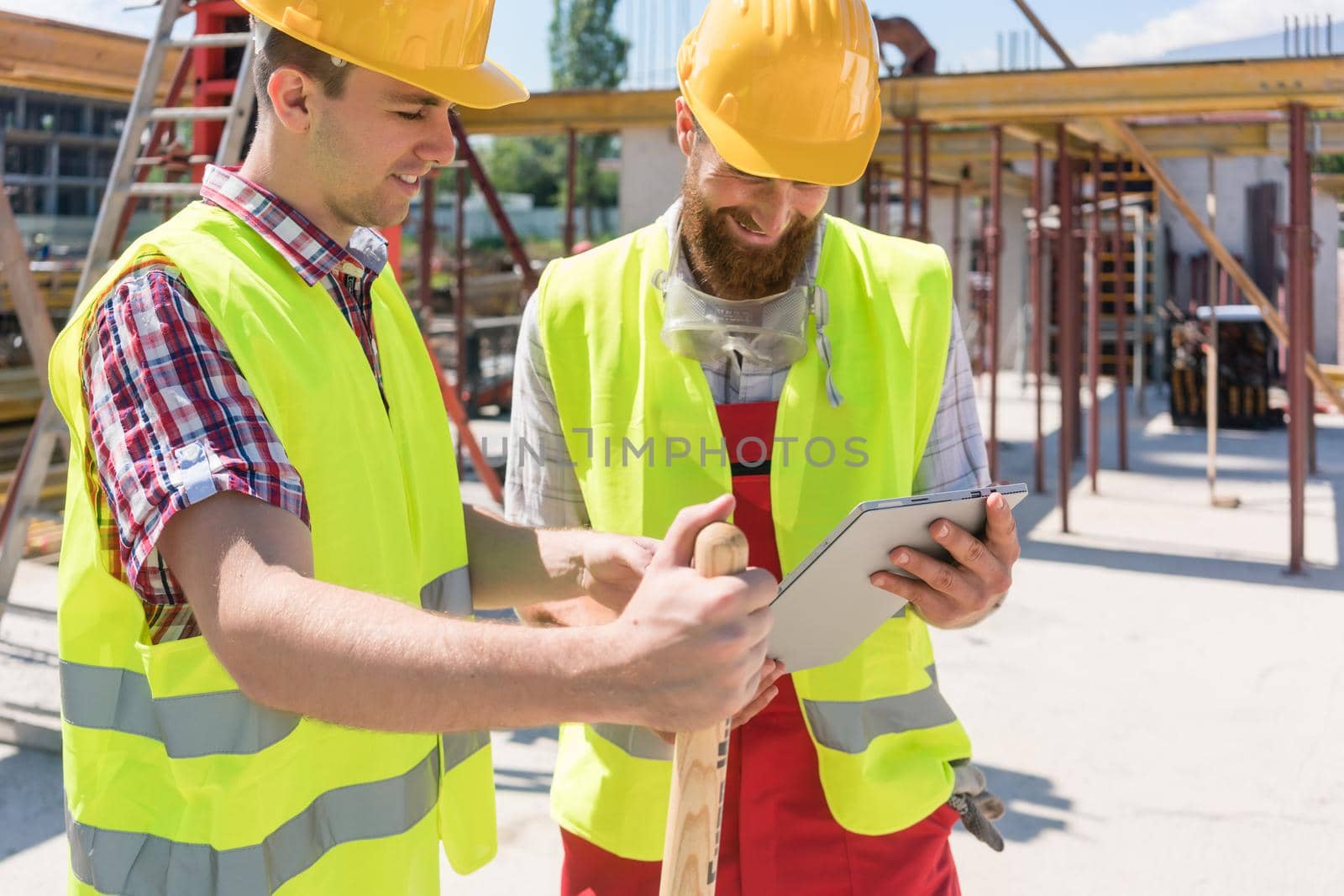 Two young construction workers smiling while using a tablet for online communication through social media during break at work