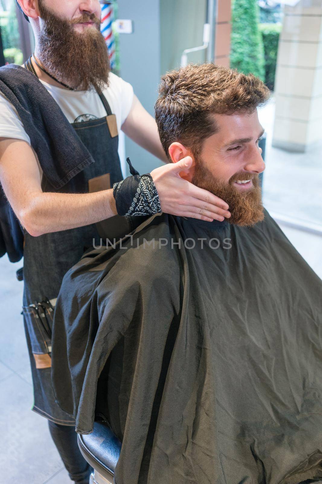 Handsome young man sitting on hair styling chair, while smiling and thinking of a different shape for his beard at the advice of his barber in the beauty salon
