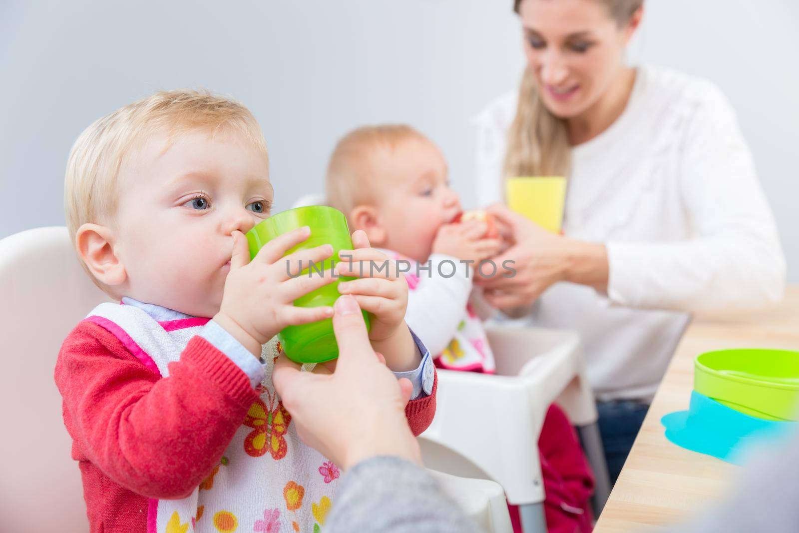 Portrait of a cute baby boy learning to drink water from a plastic glass, while sitting in a high chair next to another baby at home or in a modern daycare center for infants