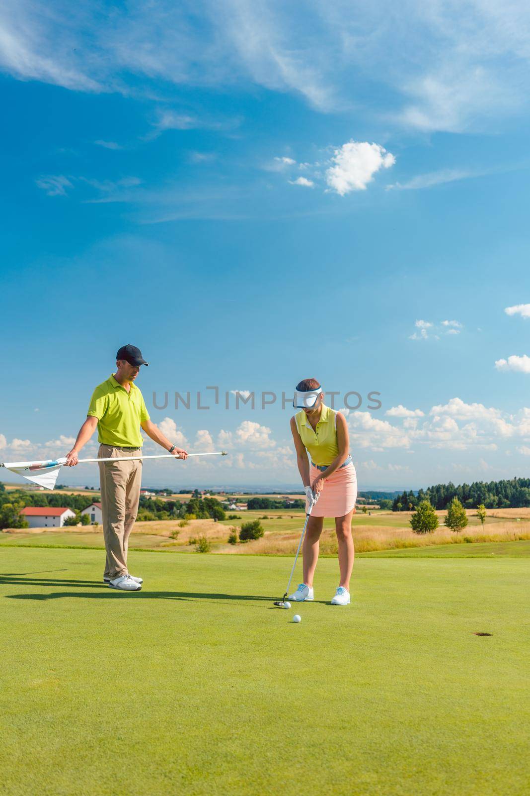 Full length of a woman calculating the trajectory of the ball to the hole, while playing professional golf with her male match partner or instructor outdoors in summer