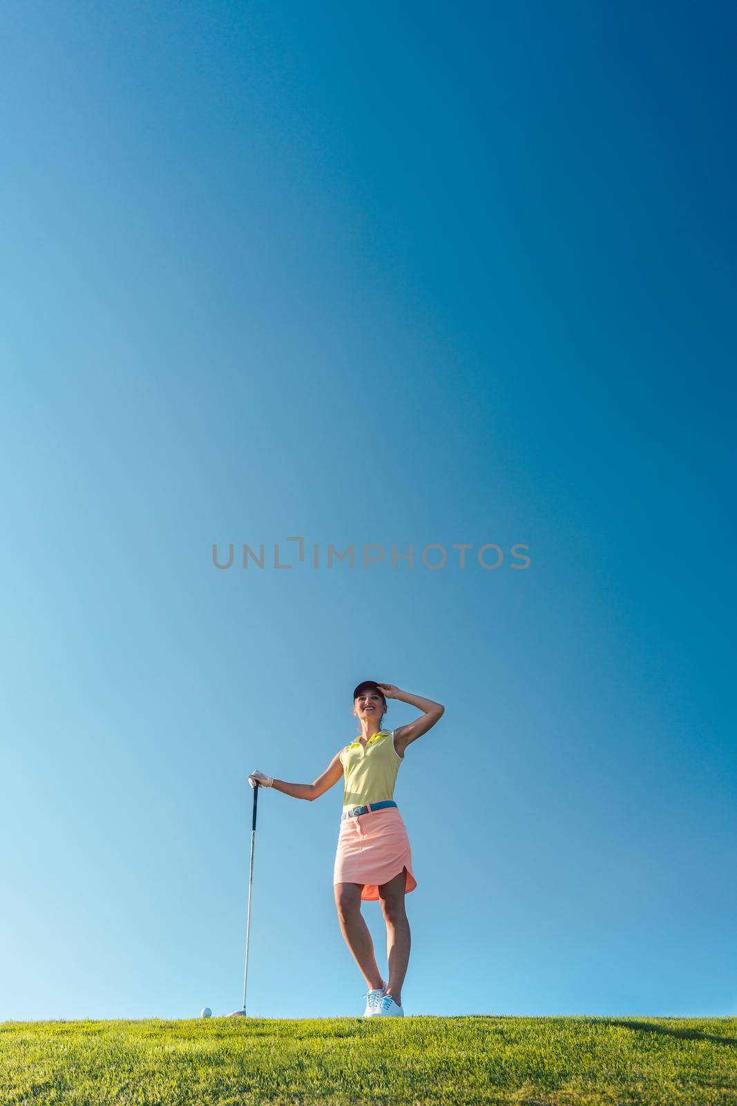 Low-angle view of a fit and cheerful woman wearing golf trendy outfits, while looking away during practice on the green grass of a professional golf course