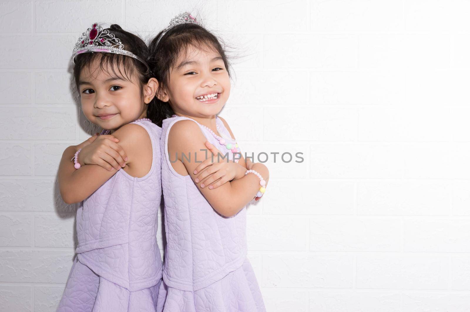 Female asian identical twins sitting on chair with white background. Wearing purple dress and accessories. Standing back to back with each other and having fun