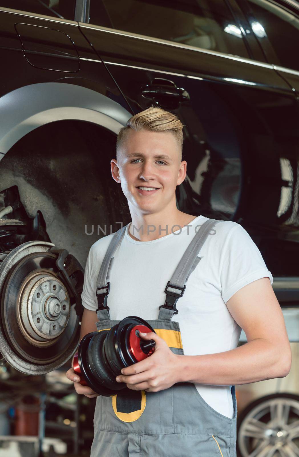 Low-angle view portrait of a confident young auto mechanic holding a new air suspension system, while working at the tuning of a car in a modern automobile repair shop