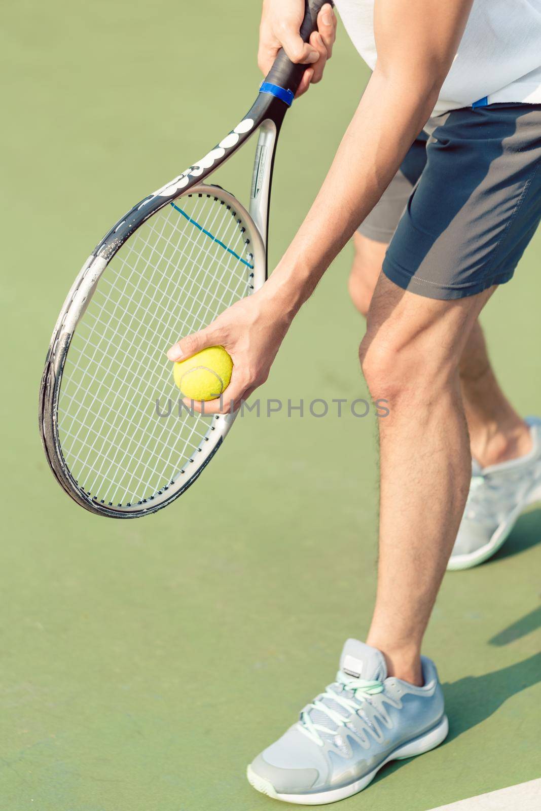 Low section of a professional player wearing gray sport shoes while holding the ball and the tennis racket during match on green surface