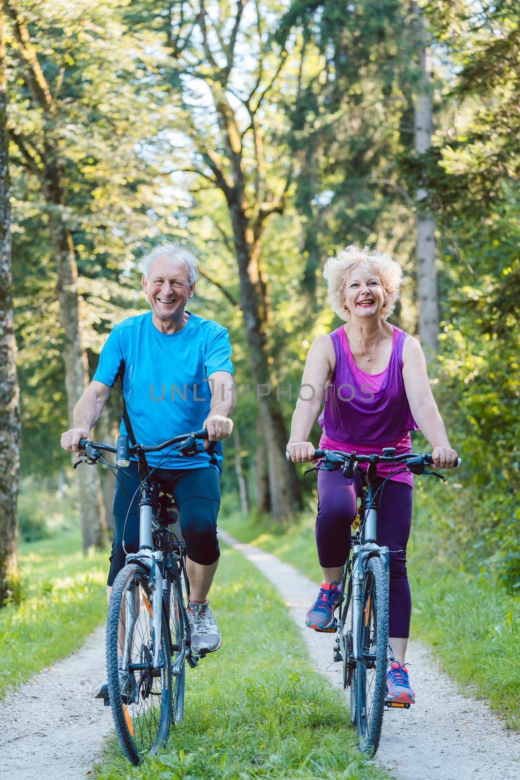 Full length of a happy and active senior couple wearing cool fitness outfits while riding bicycles outdoors in the park