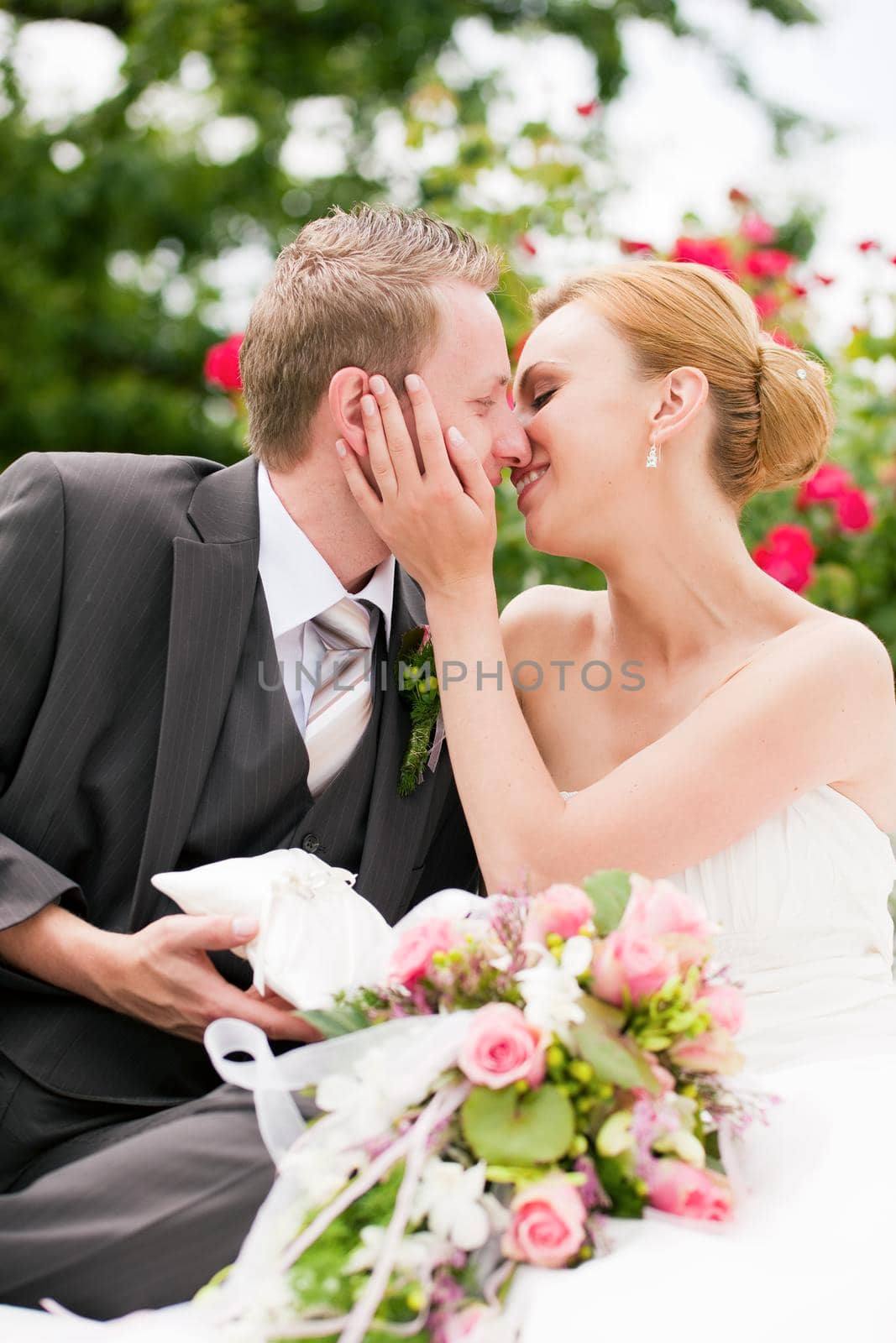 Wedding - groom kissing the bride in a park, he is holding the rings, roses in the background