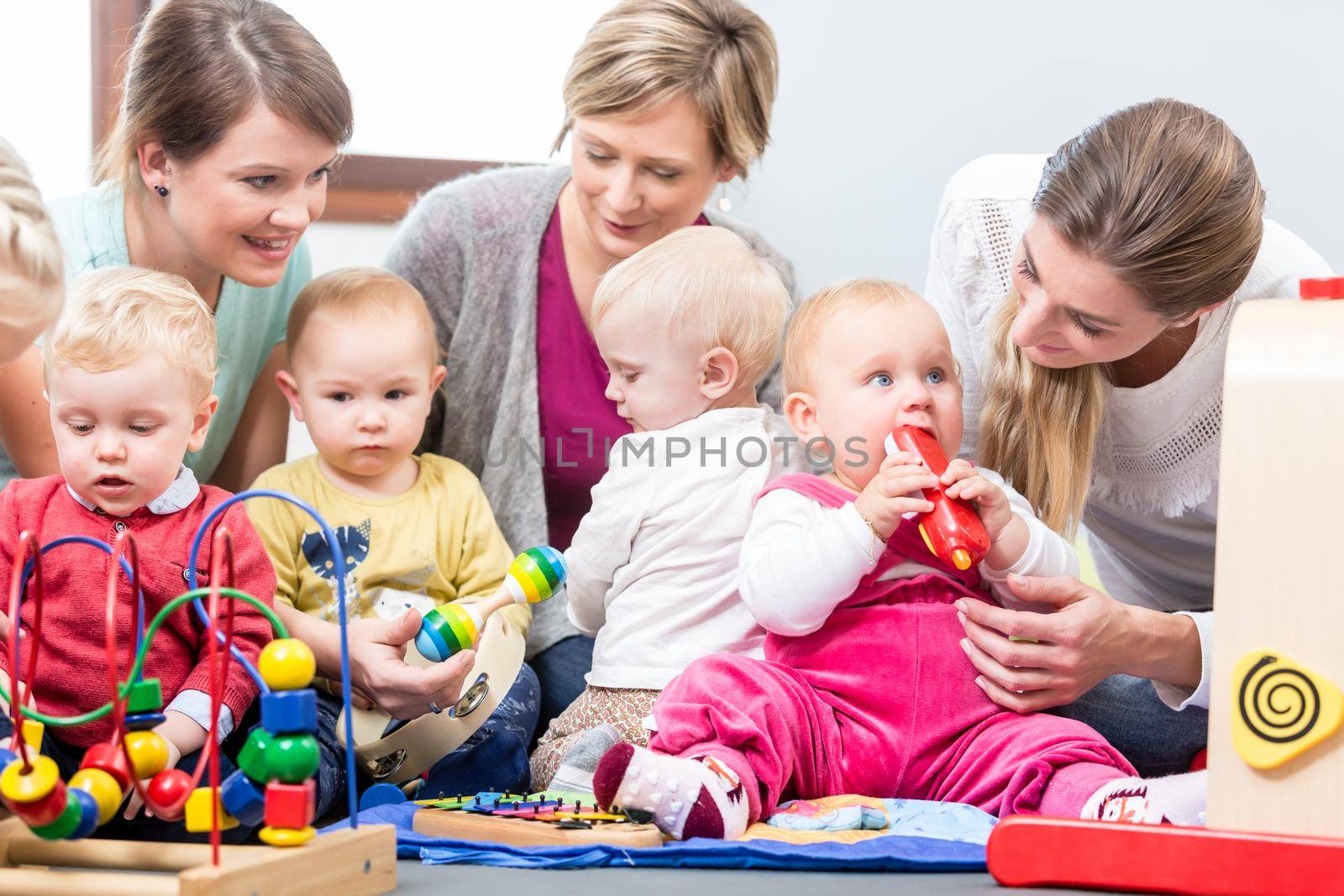 Three dedicated and happy young mothers sitting together on the floor, while watching their babies playing with safe multicolored toys at a modern playground for infants