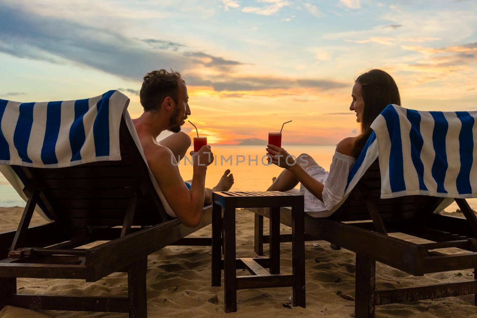Rear view of a young romantic couple in love sitting on wooden chairs, while drinking cocktails on a tropical beach at sunset during vacation or honeymoon in Indonesia