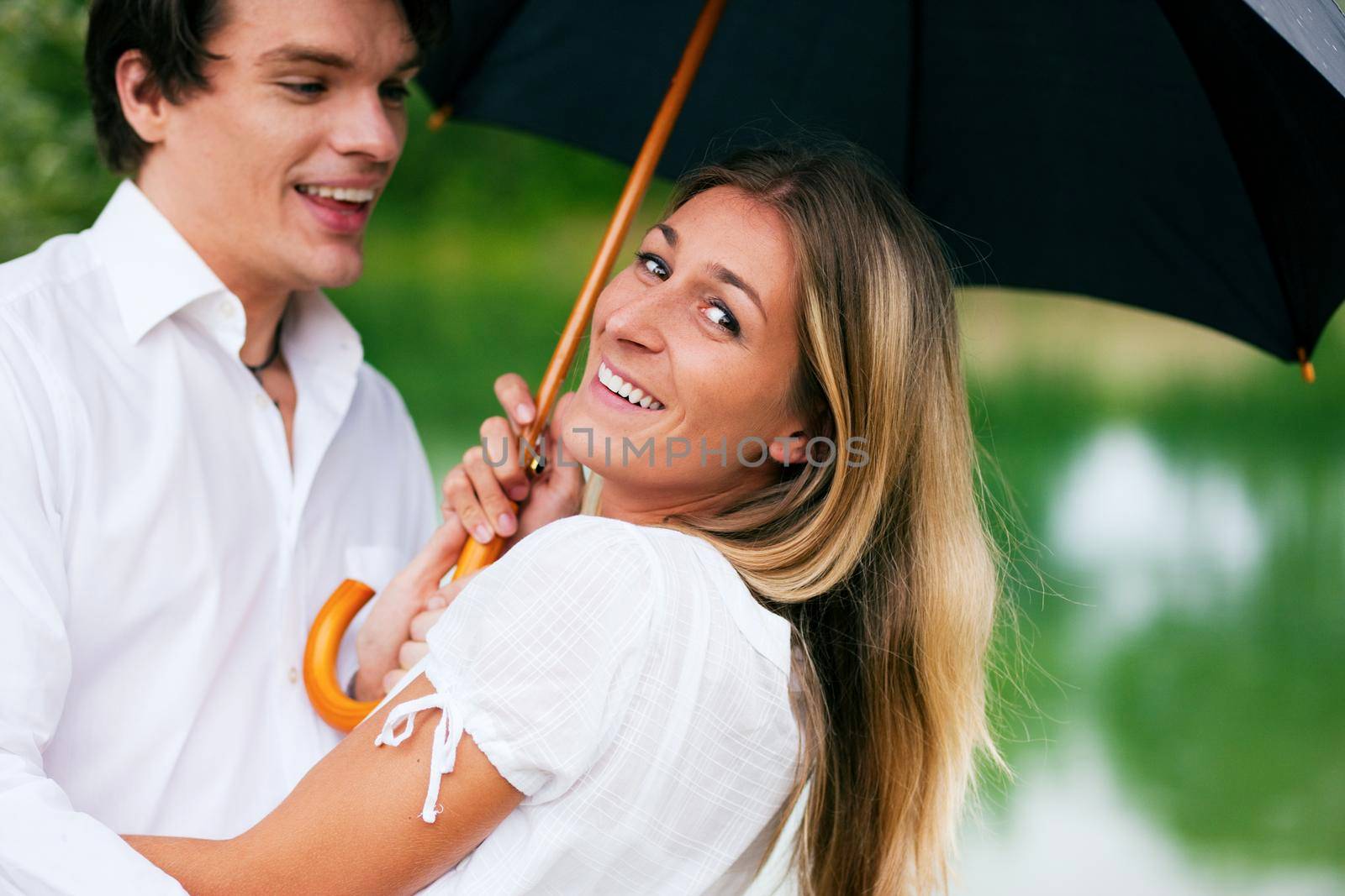 Couple (man and woman) at a lake in the summer rain with an umbrella, laughing and having fun despite of the bad weather