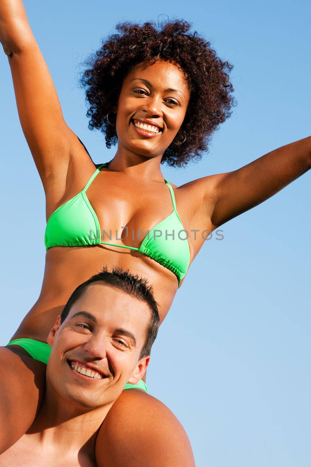 Couple in love - Woman of color in bikini sitting on her man’s shoulders under blue sky - summer and fun