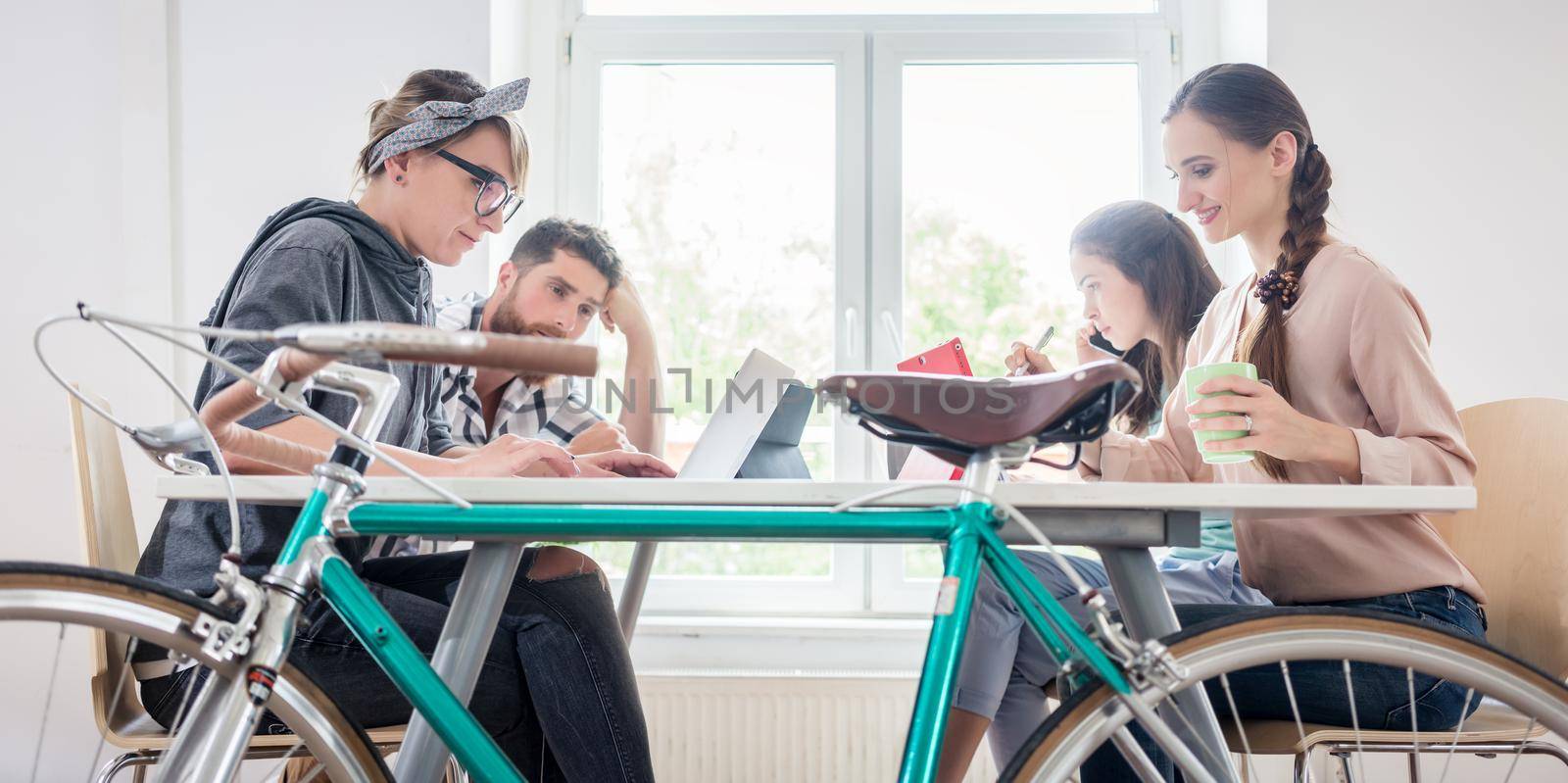 Four proficient freelancers and independent contractors co-working on tablets and laptops, connected to internet at a shared desk behind a commuter bike in a modern work hub