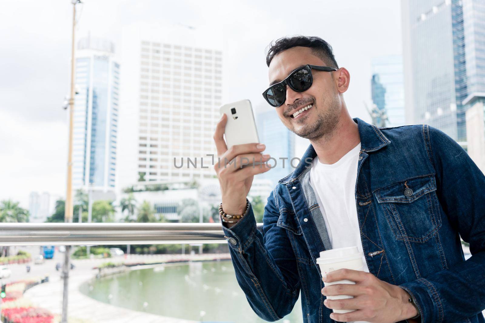 Portrait of a trendy young man smiling during online communication on his smartphone, outdoors while visiting a modern city