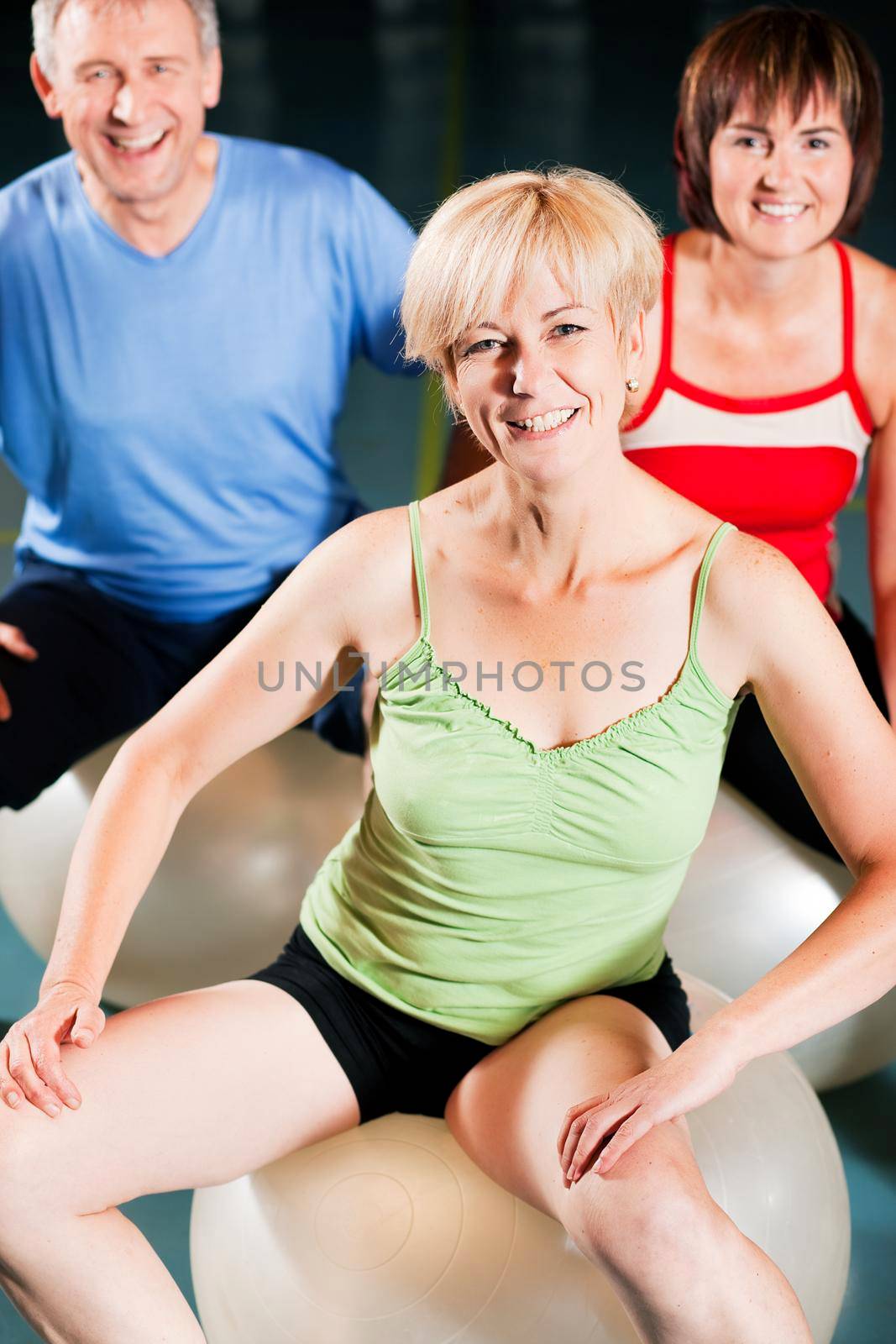 Three people - mature and senior - in the gym doing gymnastics on an exercise ball