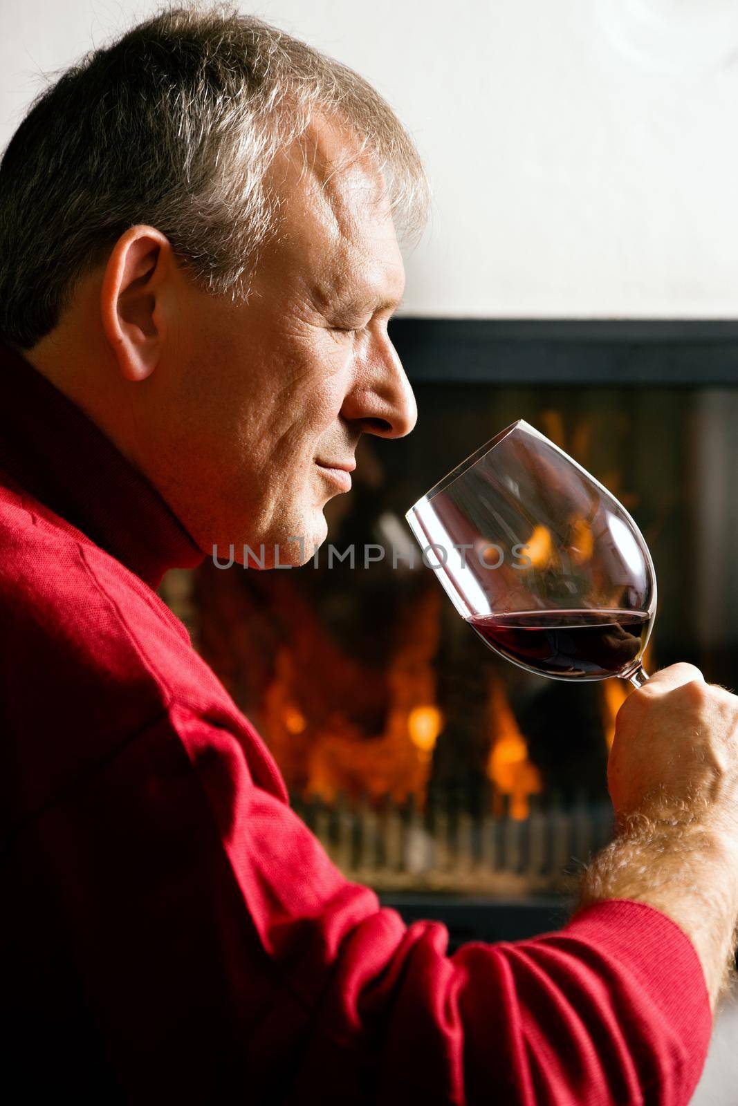 Mature man drinking a glass of read wine in front of his fireplace