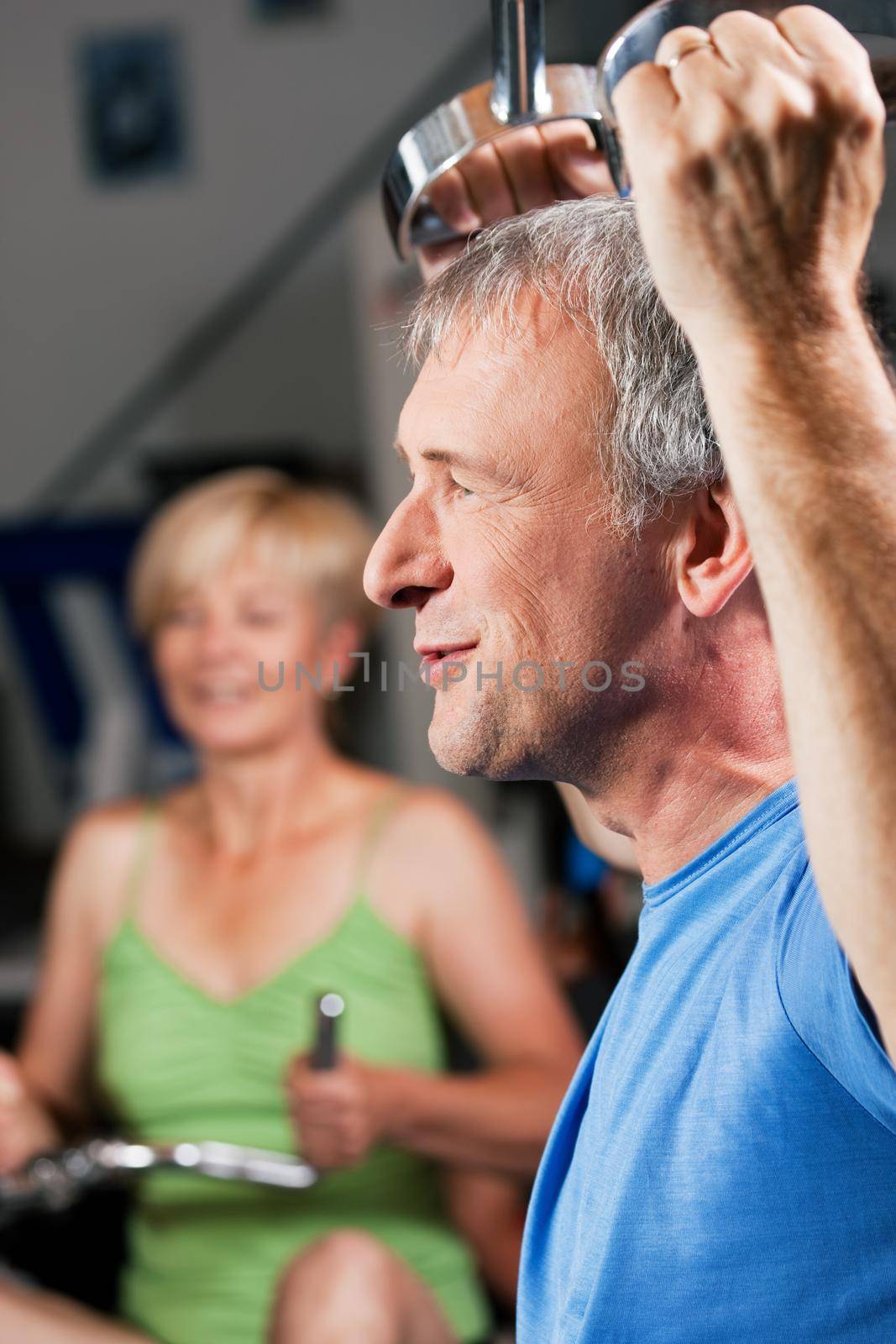 Senior couple - man and woman - in the gym lifting weights on a lat pull machine, exercising