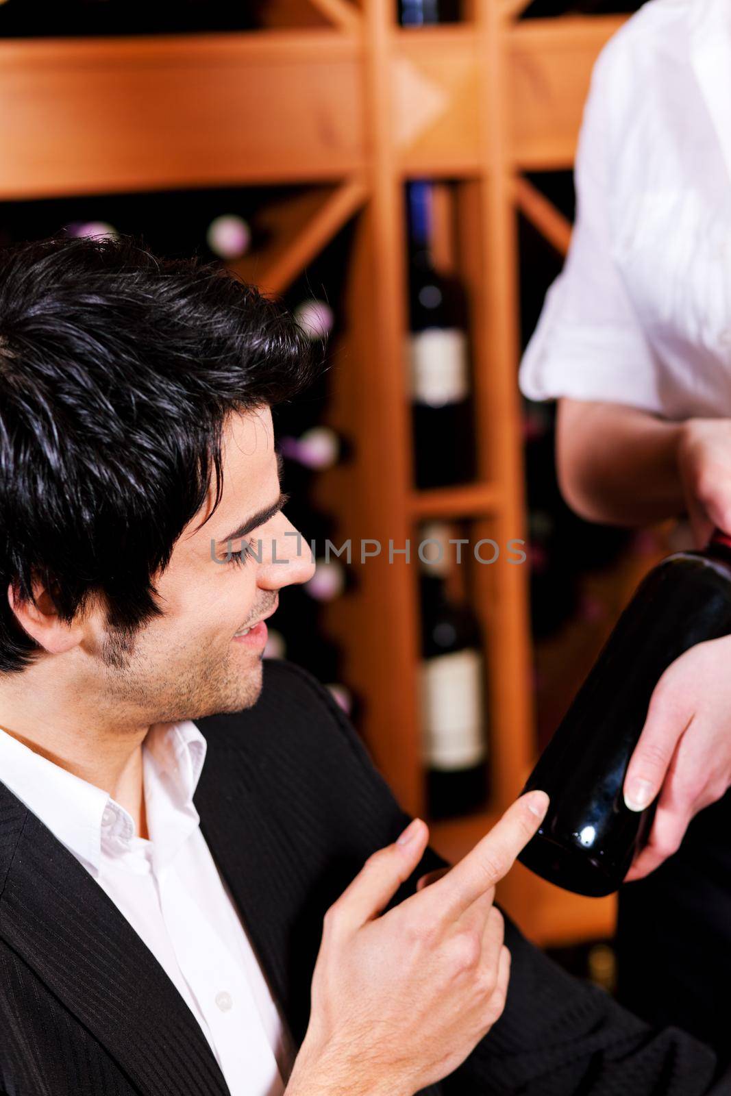 Pretty waitress in a wine bar offers a bottle of red wine to taste it a young mediterranean looking man.