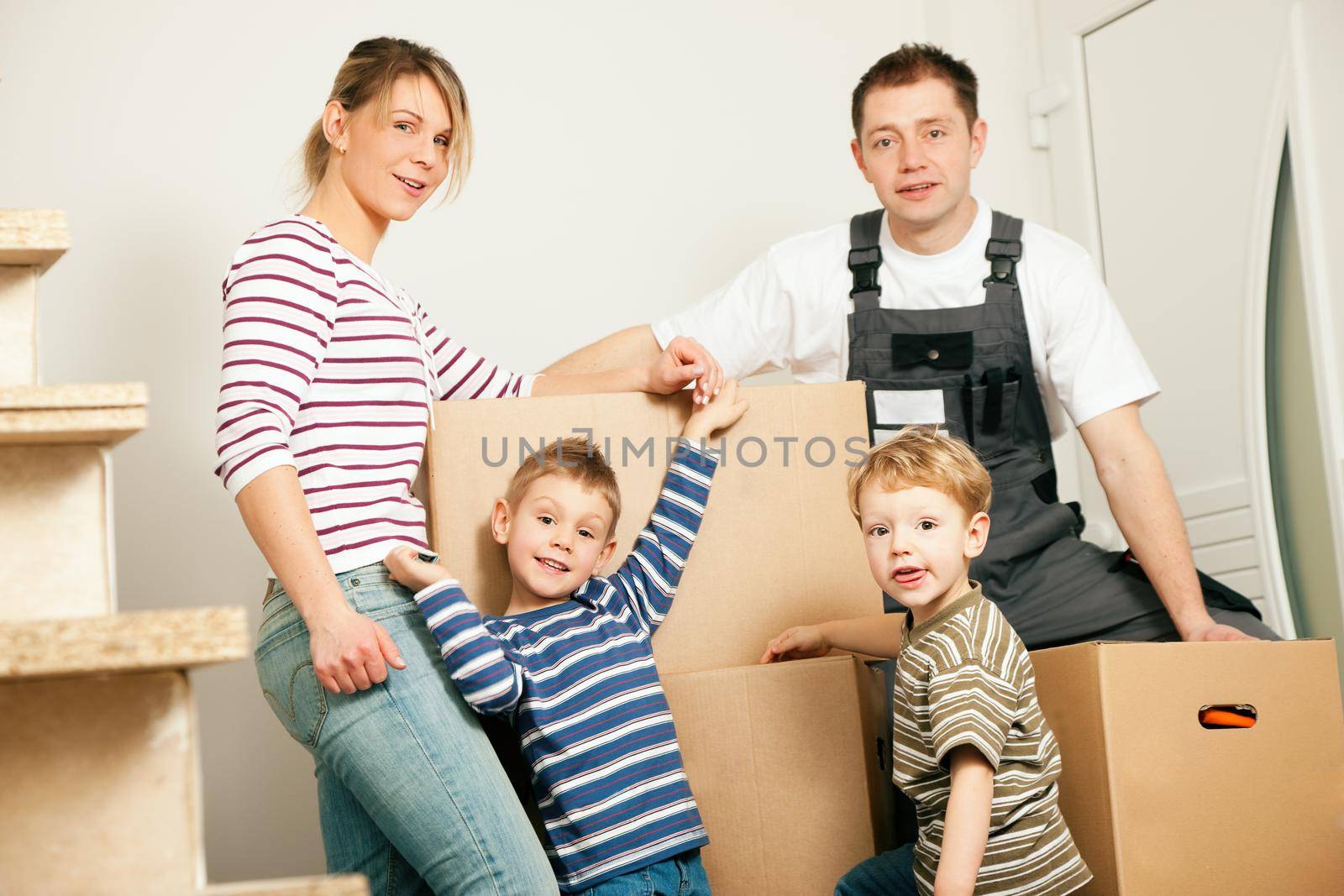 Family moving in their new house. They are standing in front of a stack of moving boxes being happy. Father is dressed in a way that can also represent a mover