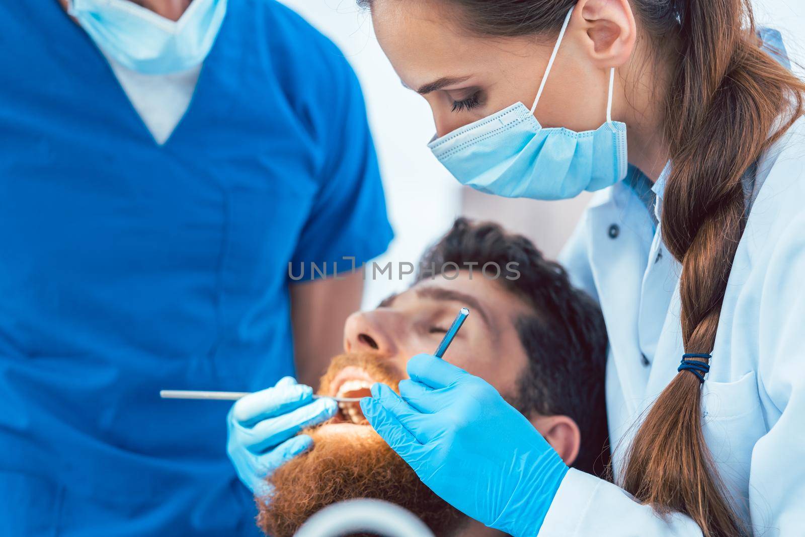 Side view of a reliable female dentist using sterile instruments and blue surgical gloves, while cleaning the teeth of a patient in the dental office of a modern clinic