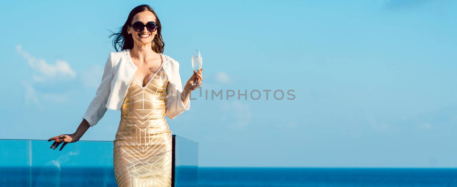 Woman drinking sparkling wine looking over ocean wearing an expensive dress