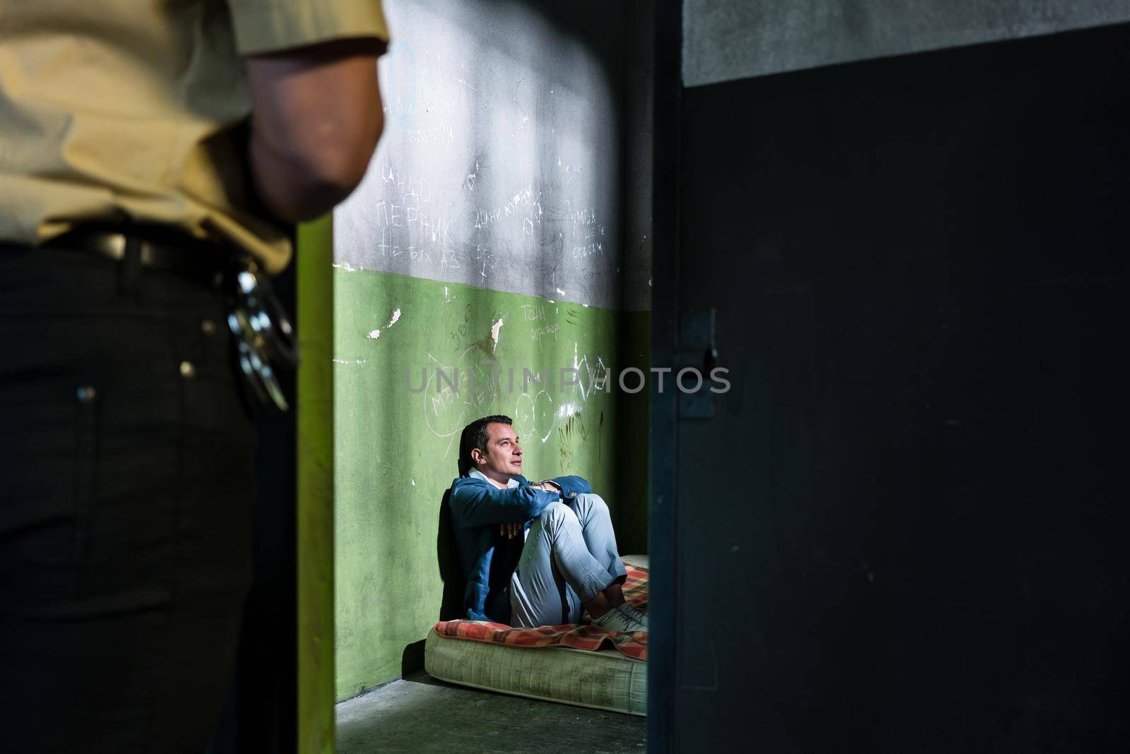 Side view of a young male prisoner thinking while sitting alone on a dirty mattress in an obsolete prison cell guarded by a police officer