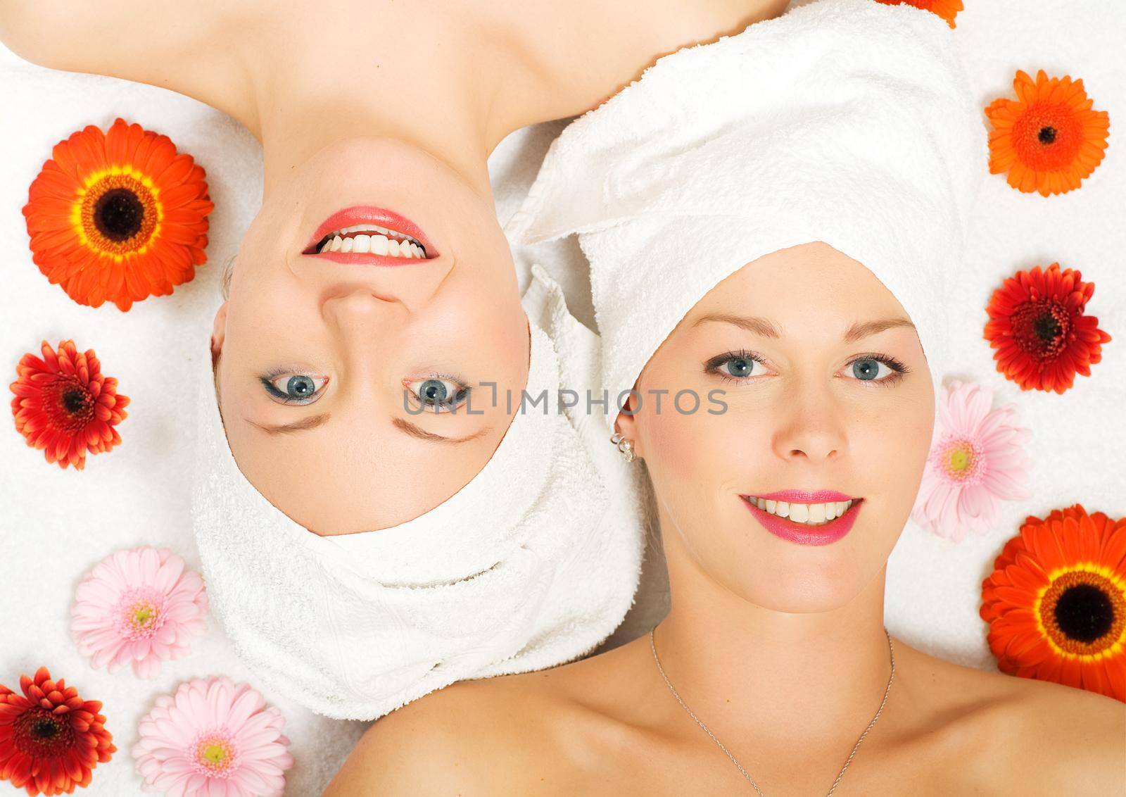 Two girls relaxing in a wellness set-up seen from above, vertically aligned, with some flowers, open eyes, smiling