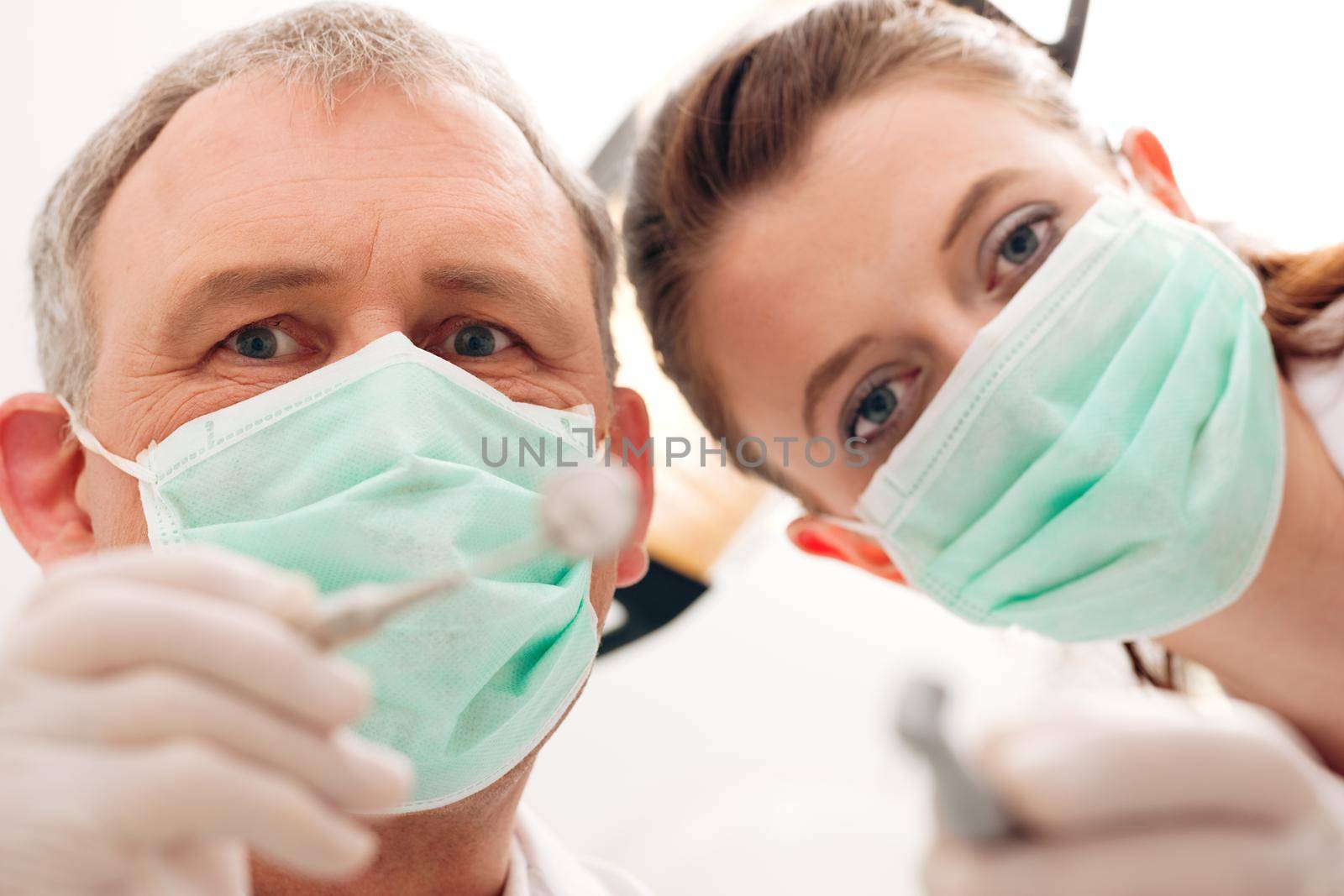 Dental treatment with dentist and dental assistant bowing over the patient as seen from patient's perspective. They have drills and angled mirrors, focus on eyes of dentist