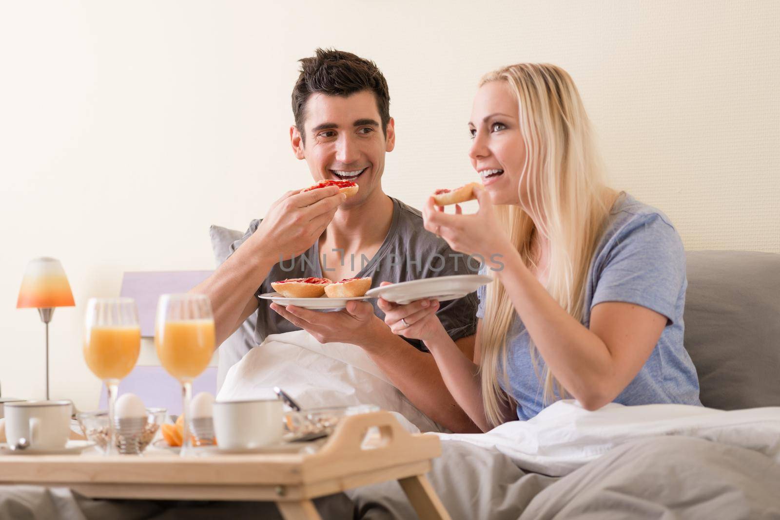 Young couple celebrating with breakfast in bed sitting eating berry jam o toast accompanied by fresh orange juice and boiled eggs