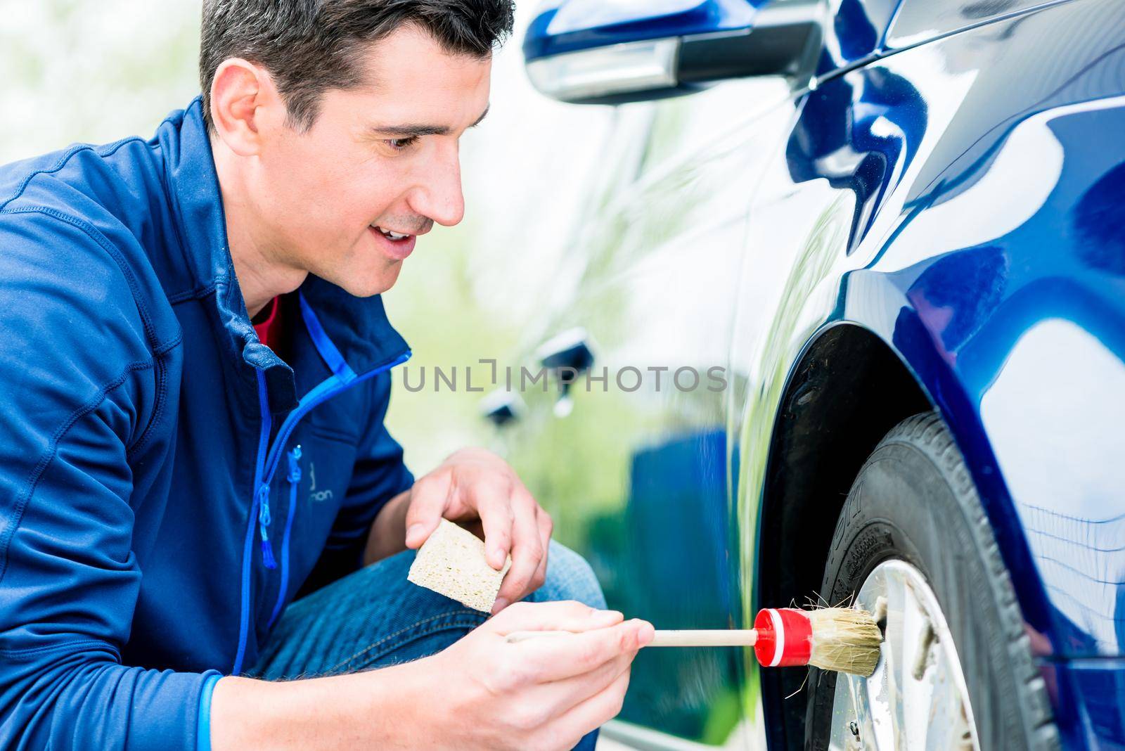 Smiling attractive man cleaning the alloy hubs on his car tyres using a soft bristle brush and soap