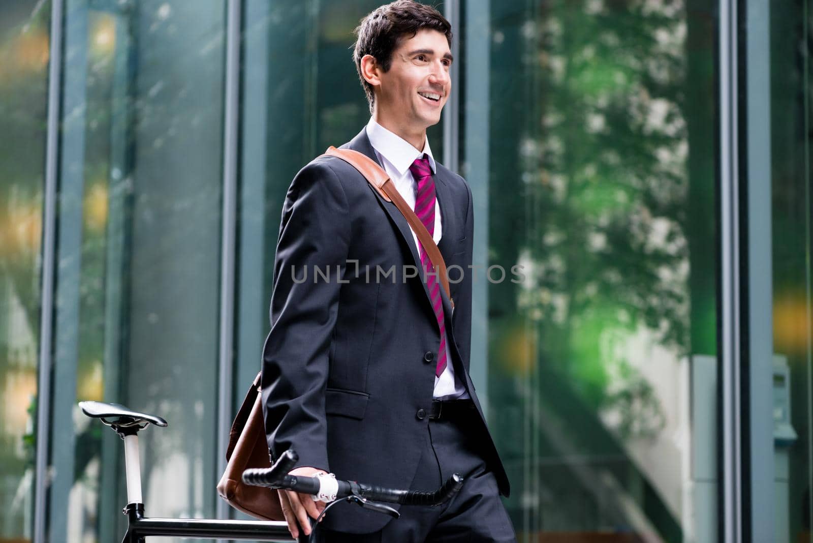 Smiling handsome young business man walking through town with his bike wheeling it along a pedestrian walkway as he commutes to work