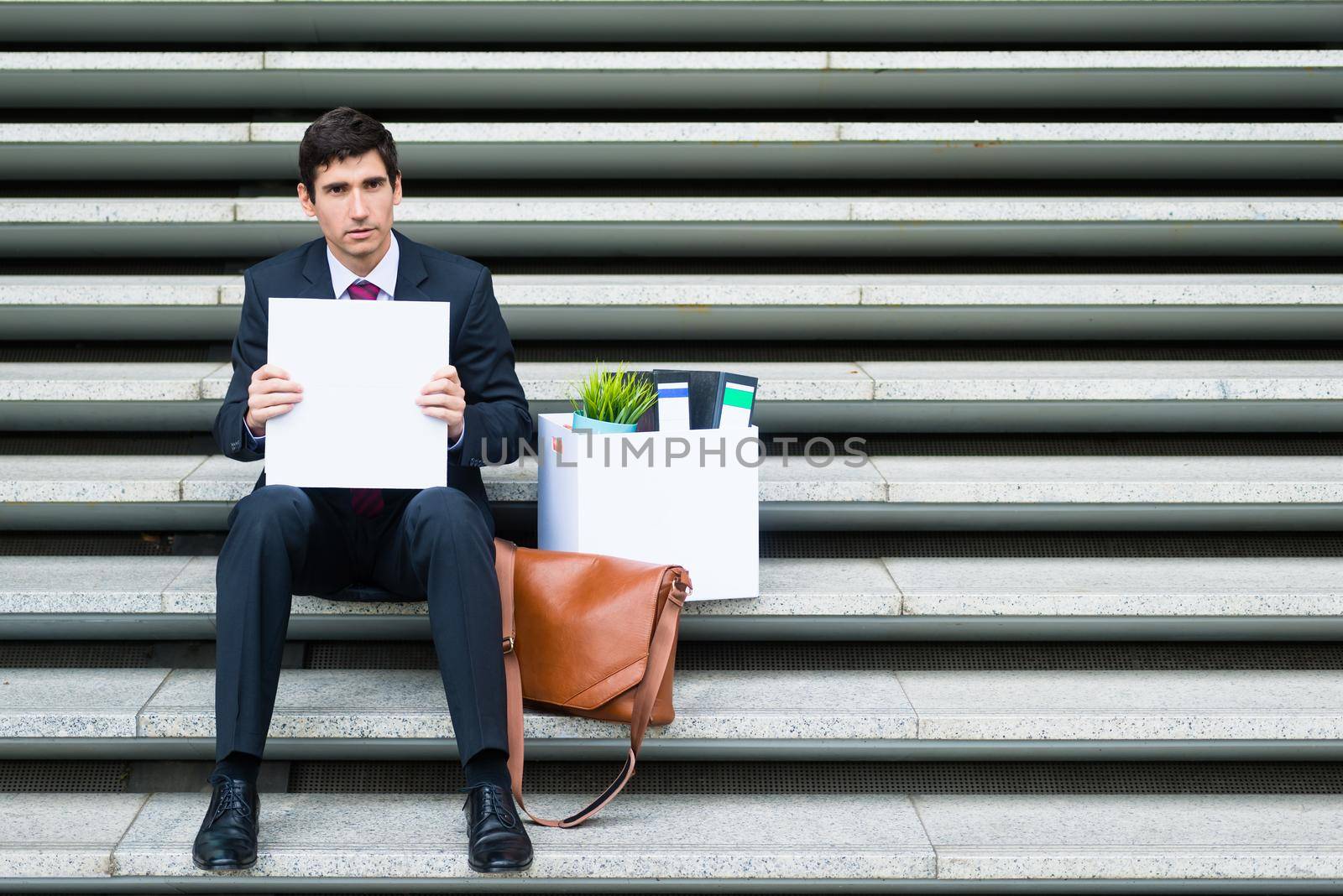 Disconsolate businessman holding a blank sign as he sits on a flight of steps in town with his briefcase and office files after being made redundant
