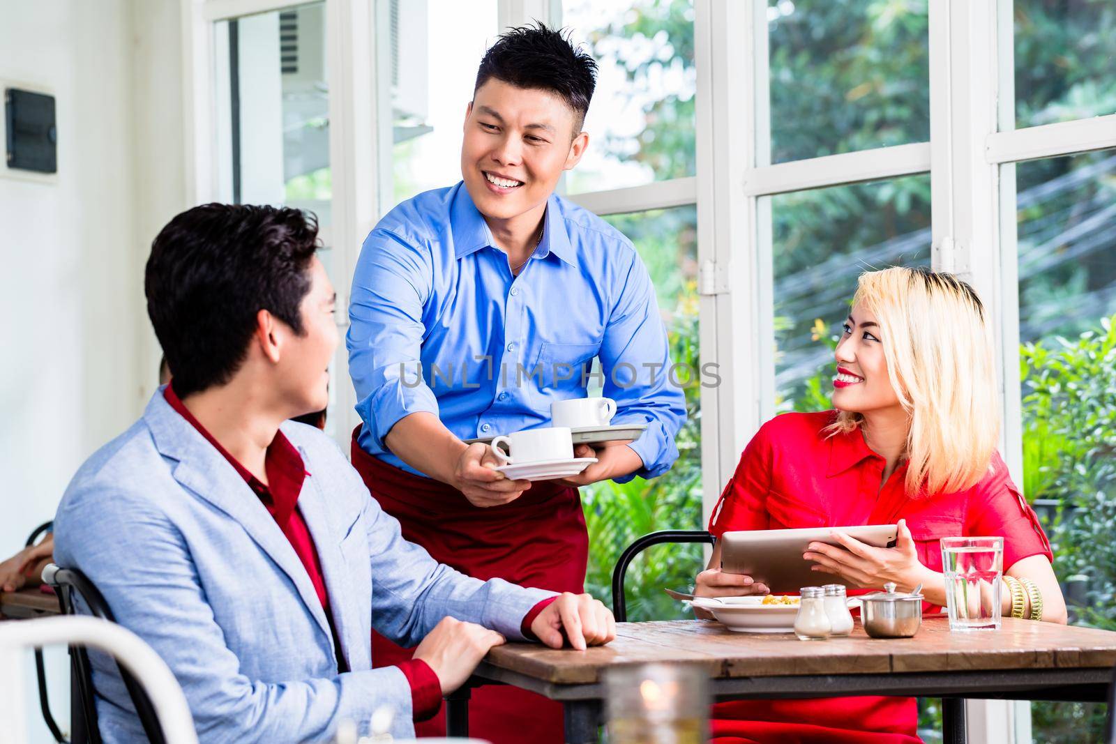 Happy Asian waiter serving a stylish couple coffee as they sit at a table in a restaurant enjoying a meal