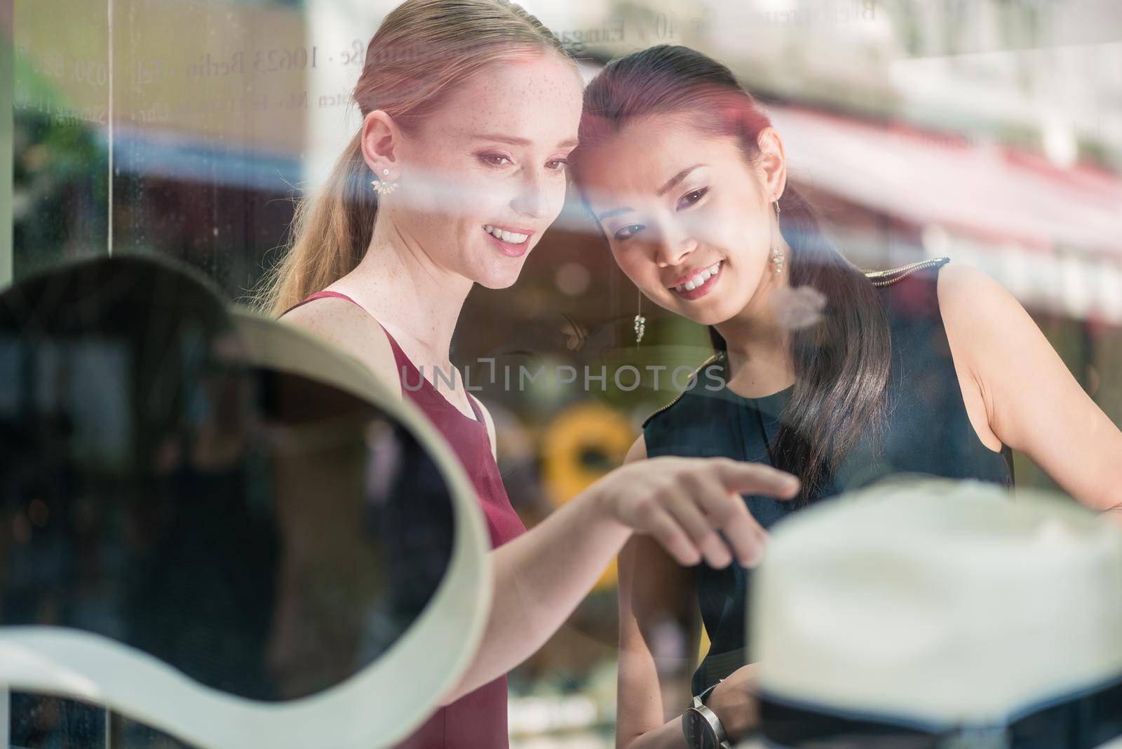 Two women friends shopping in a fashion boutique pointing to a display of trendy hat viewed through the glass with reflections