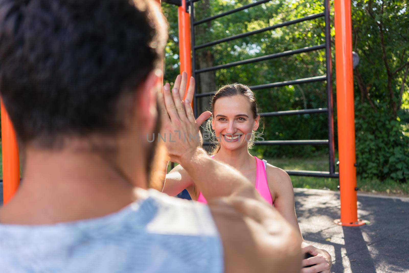 Over the shoulder view of a cheerful young woman giving high-five to her partner, while doing crunches outdoors during couple workout in a fitness park