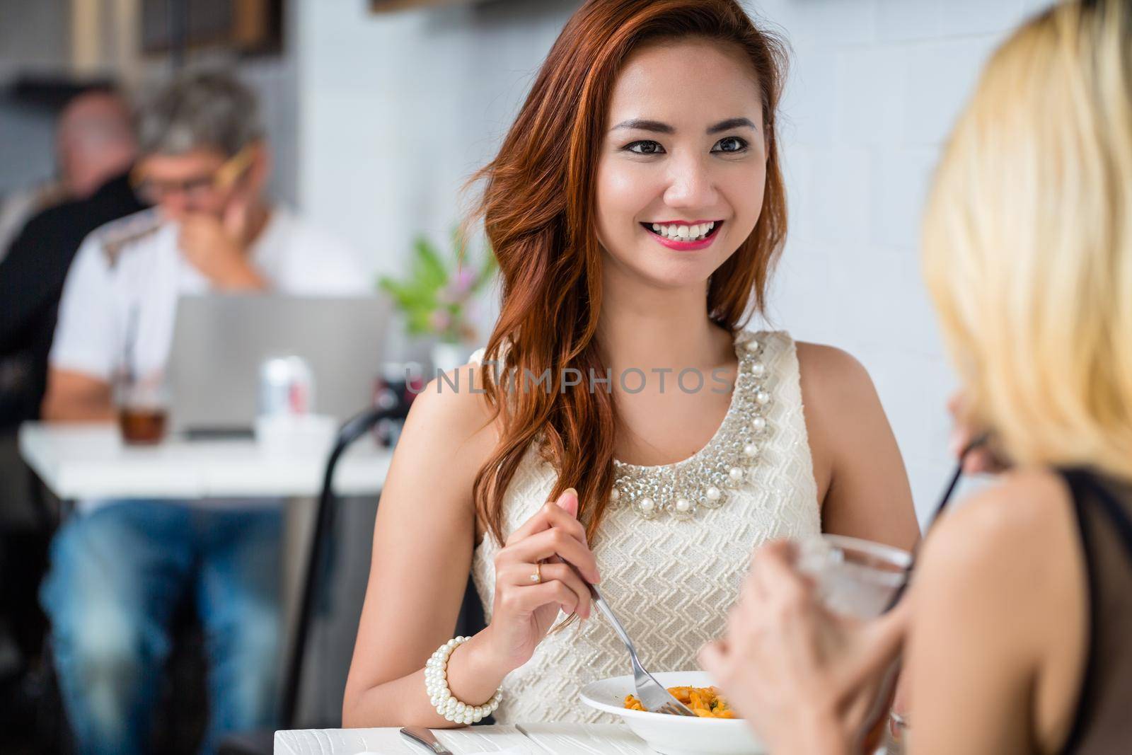 Attractive elegant young Asian woman having lunch with a friend at a restaurant sitting at the table together smiling and chatting, over the shoulder view