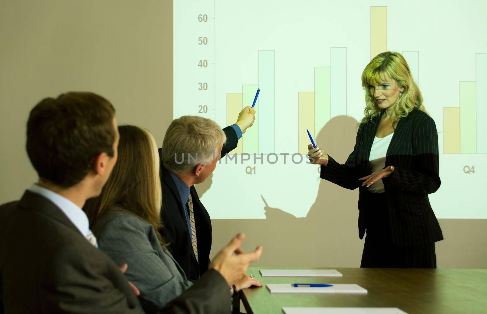 group of people listening to a video presentation held by a blond woman, one attendant asking something
