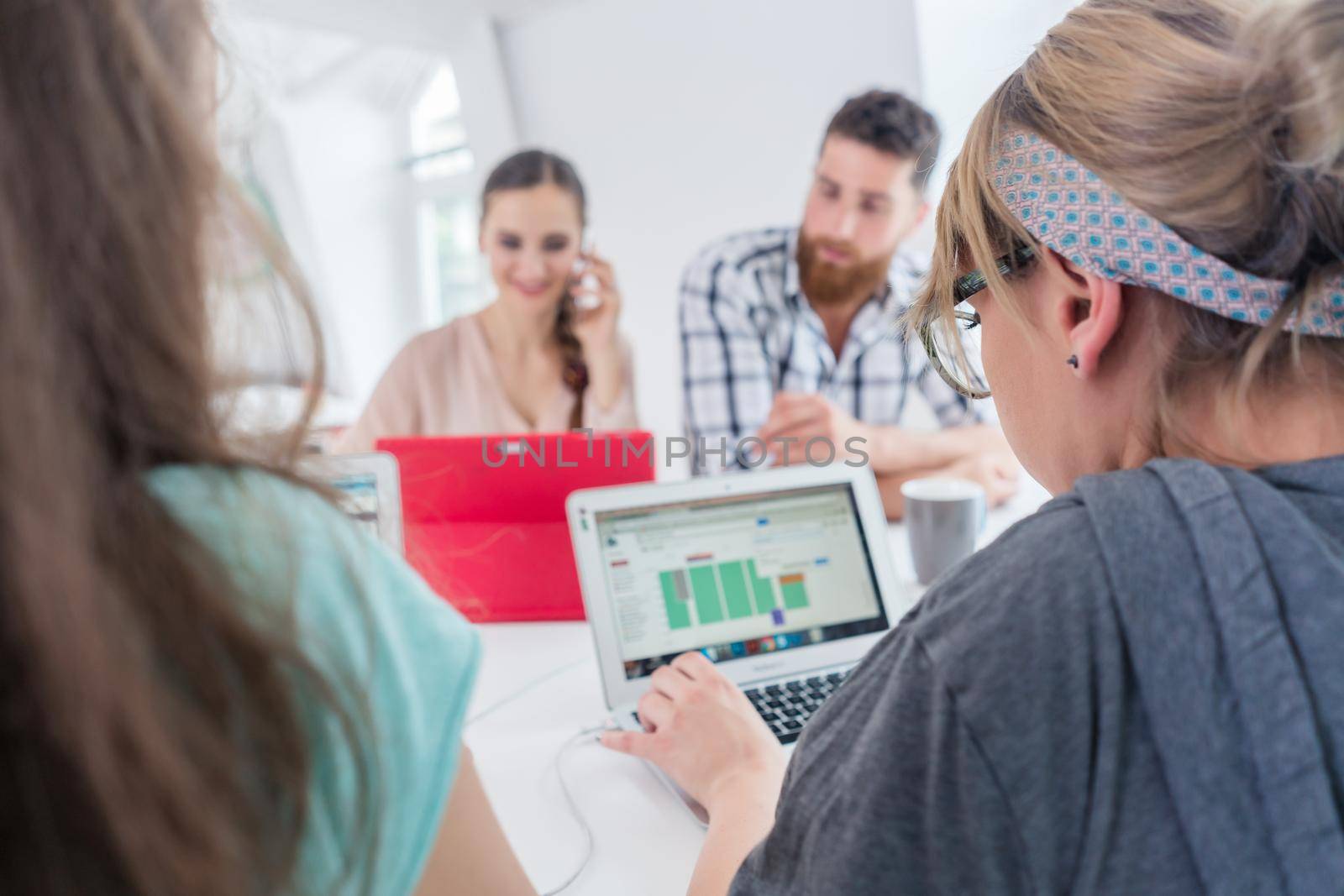 Active female entrepreneur talking on mobile phone, while using a tablet next to her helpful co-worker at a shared desk in a modern collaborative office space