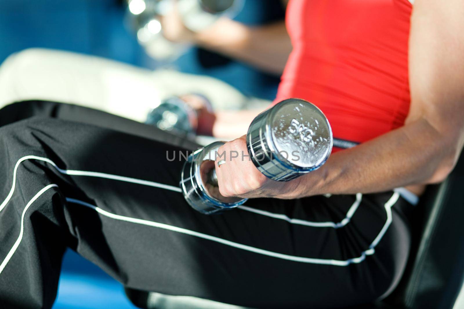 Woman lifting hand weights in a gym, in the background a man doing the same; focus on hand of woman