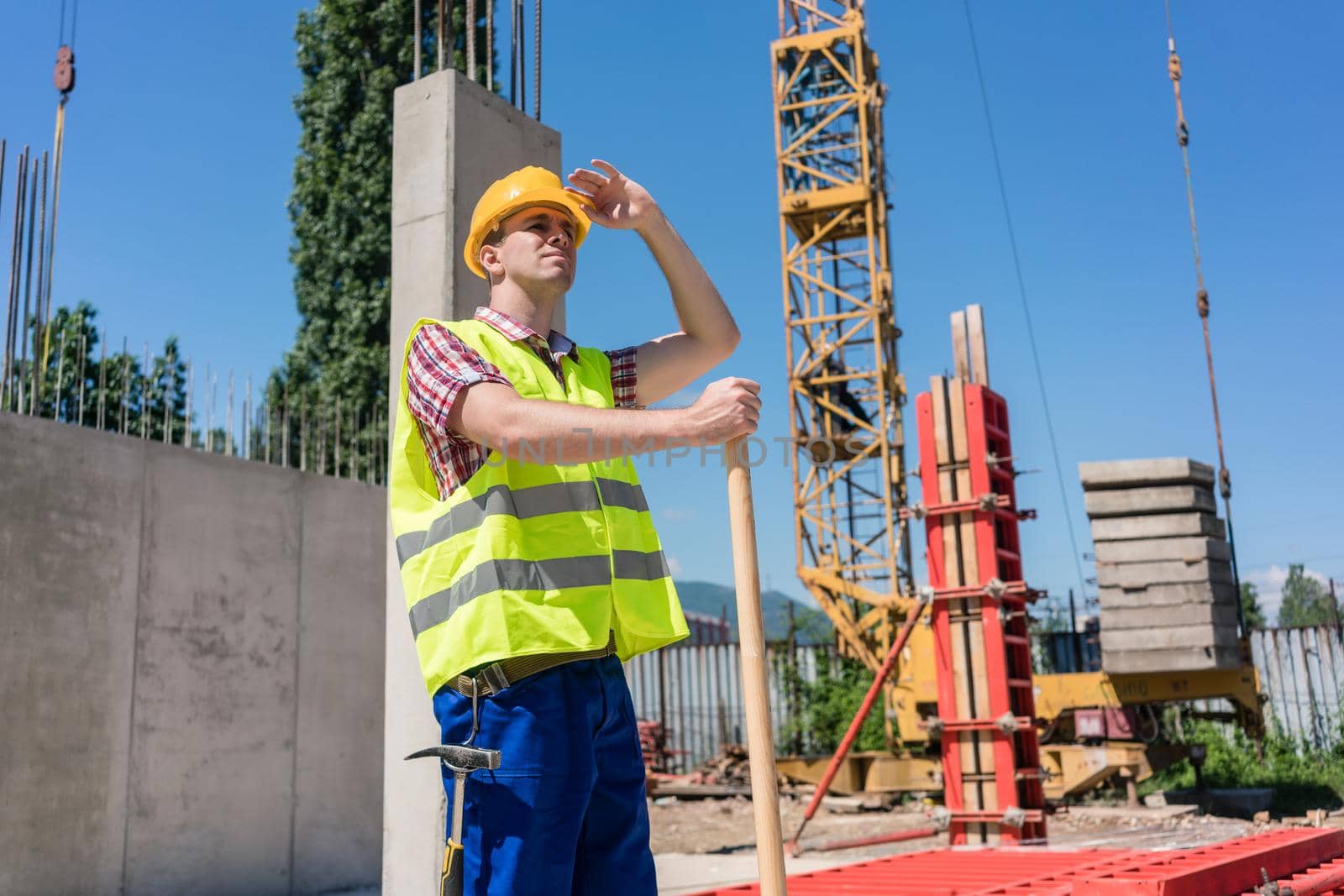 Young blue-collar worker looking up with a worried facial expression while wearing safety equipment during work on the construction site in a sunny day