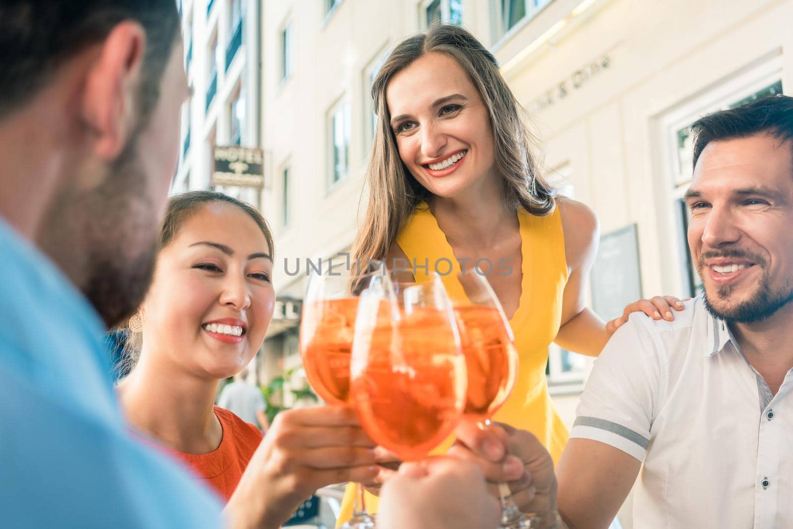 Low-angle view of a multi-ethnic group of four friends celebrating together with an orange refreshing summer drink at a trendy restaurant downtown