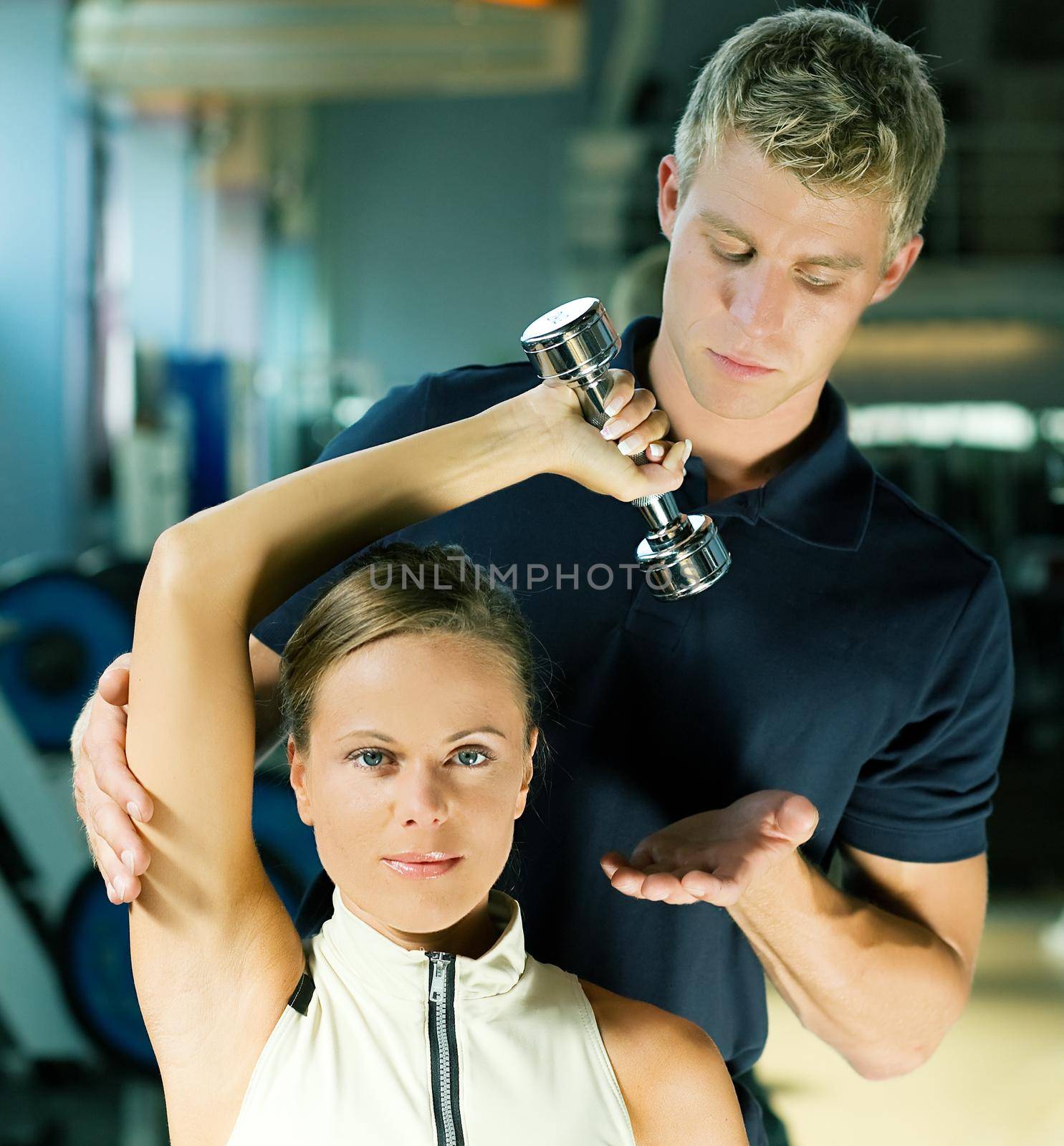 Young woman lifting a dumb-bell in the gym assisted by her personal trainer